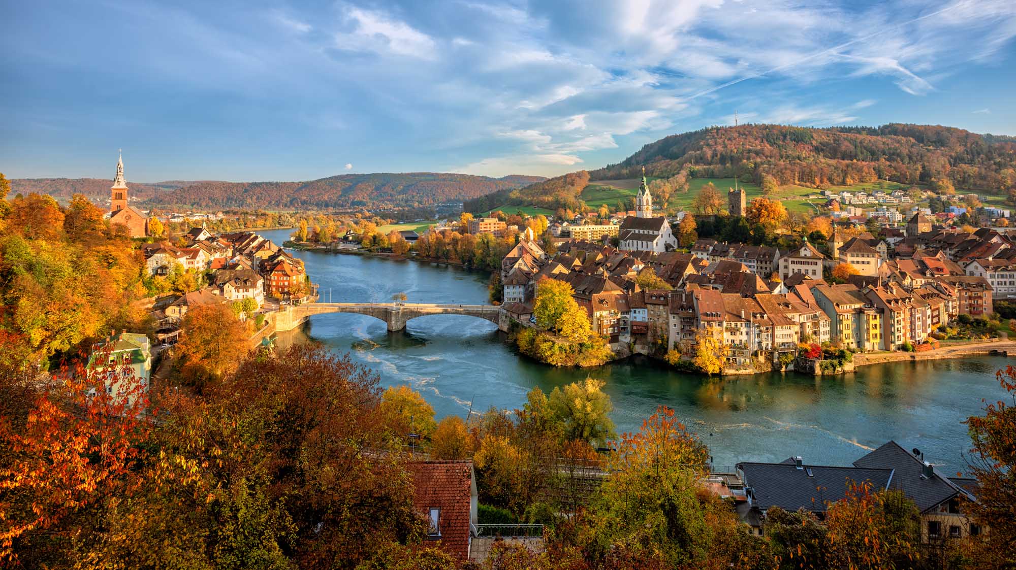 Laufenburg Old Town on the Rhine River A picturesque autumn view of Laufenburg's historic old town straddling the Rhine River on the Swiss-German border, featuring traditional architecture, a stone bridge, church spires, and colorful fall foliage reflected in the blue waters below, with forested hills rising in the background.