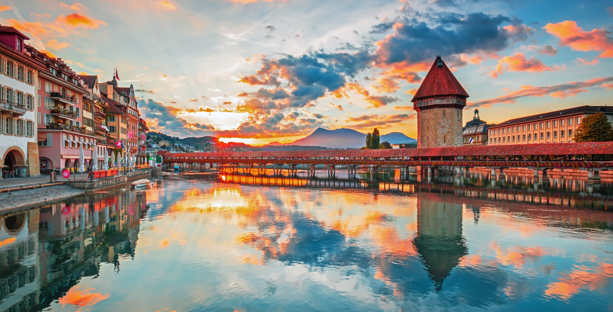 A stunning sunset view of Lucerne, Switzerland, featuring the iconic wooden Chapel Bridge (Kapellbrücke) with its distinctive red roof tower, colorful medieval buildings reflected in the calm waters of Lake Lucerne, and snow-capped mountains silhouetted against a vibrant golden and blue sky.