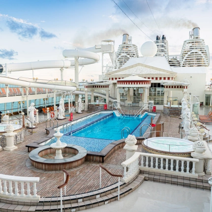 Luxurious Pool Deck on Modern Cruise Ship An expansive cruise ship pool deck featuring a large turquoise swimming pool, hot tubs, water slide, and elegant white architectural elements. The deck is populated with guests enjoying various amenities, with distinctive curved residential towers visible in the background against a partly cloudy sky.