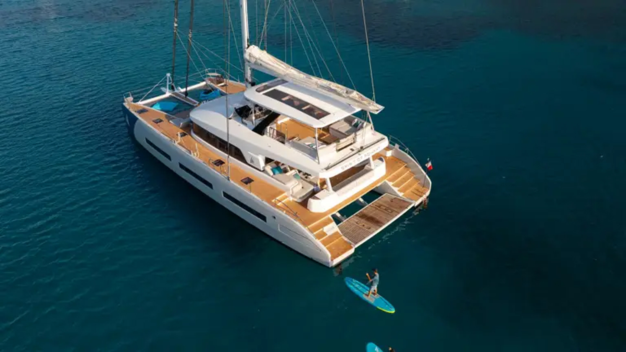 Luxury Catamaran Yacht with Water Sports A modern white and natural wood luxury catamaran yacht anchored in stunning turquoise waters, with passengers enjoying water activities including paddleboarding and swimming in the crystal-clear Mediterranean or tropical sea.