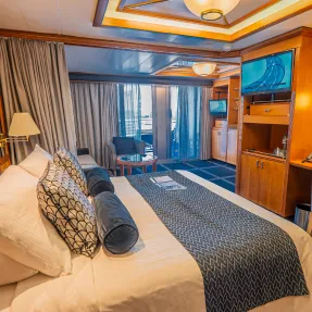 Luxury Cruise Ship Cabin with Ocean View An elegantly appointed cruise ship stateroom featuring a king bed with decorative pillows, floor-to-ceiling windows overlooking the ocean, modern wood cabinetry, warm ambient lighting, and contemporary furnishings. The cabin showcases premium cruise accommodations with sophisticated blue and orange color accents and a flat-screen television.