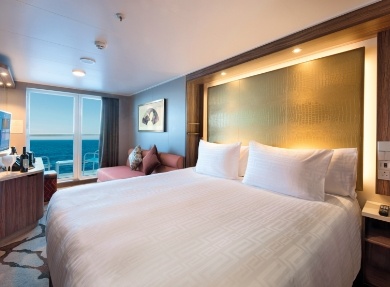 Luxury Cruise Ship Oceanview Cabin with Balcony A spacious, modern cruise ship cabin featuring a king-sized bed with premium linens, an illuminated headboard, and floor-to-ceiling windows offering a scenic ocean view with a private balcony. The room showcases contemporary design with warm lighting, elegant furnishings, and modern amenities typical of a premium suite aboard a contemporary cruise vessel.