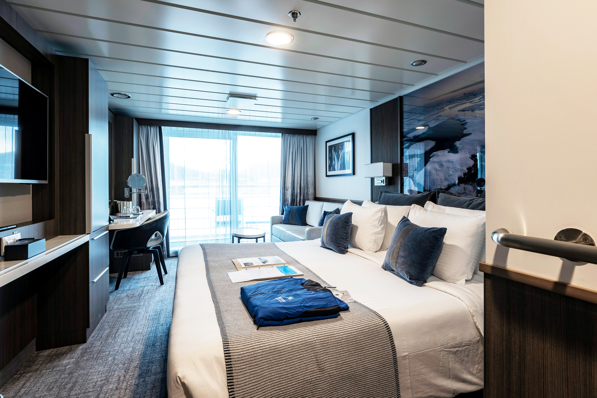 Luxury Cruise Ship Stateroom with Ocean View An elegantly designed stateroom aboard a modern cruise ship, featuring a king-size bed with premium linens, contemporary furnishings in dark wood and blue tones, a work desk, flat-screen television, and expansive balcony doors offering panoramic ocean views. The cabin exemplifies upscale cruise accommodations with sophisticated decor and modern amenities.