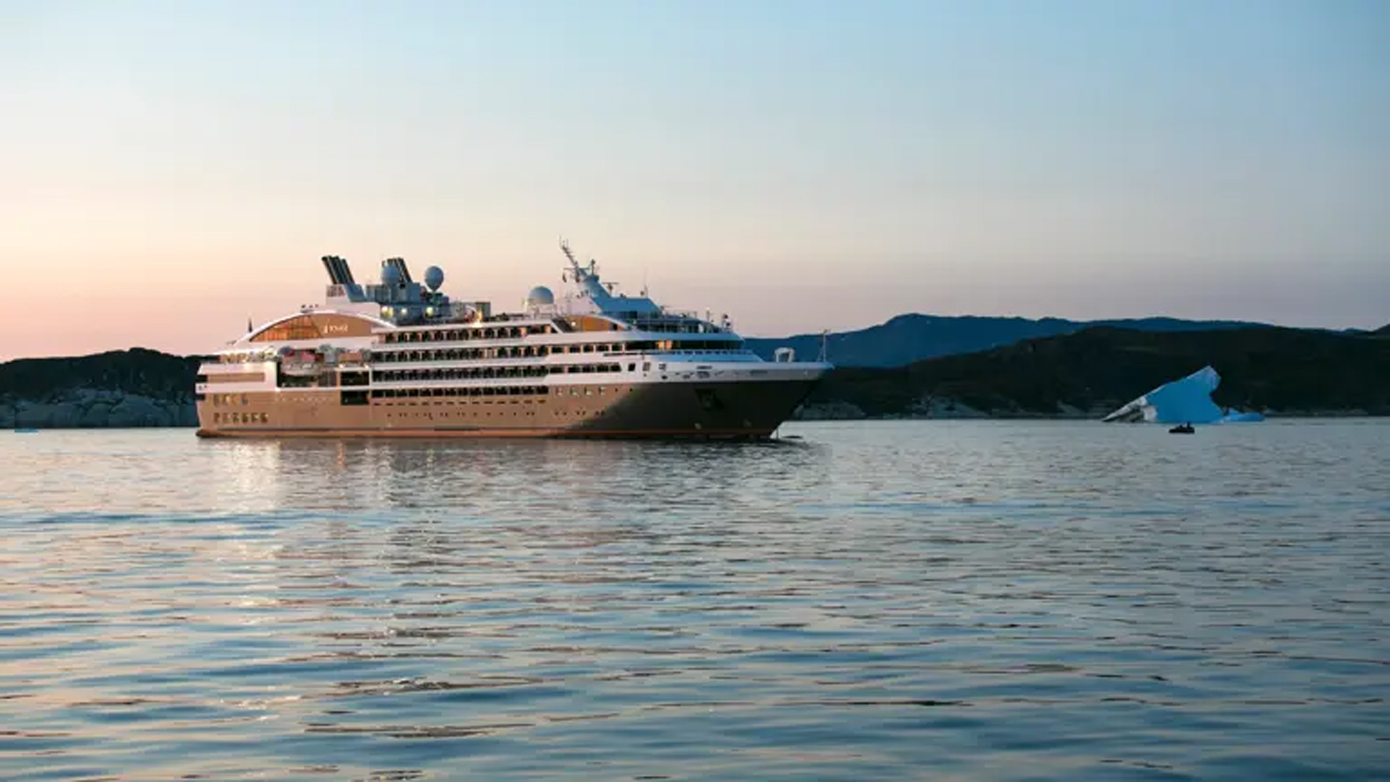 A modern expedition cruise ship anchors in calm Arctic waters near Bisko Bay, with snow-capped mountains and an iceberg visible in the background during twilight hours. The vessel features a distinctive design with multiple decks and contemporary architecture typical of luxury expedition cruising.