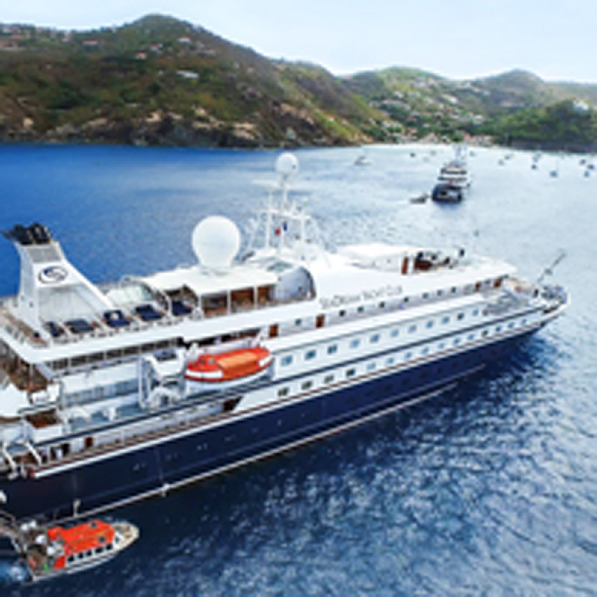 Luxury Cruise Ship in Mediterranean Bay A large white and blue luxury cruise ship anchors in a pristine Mediterranean bay surrounded by verdant hillsides with scattered coastal homes. The vessel features multiple decks, a distinctive white funnel with radar equipment, and is accompanied by smaller tender boats with red and white livery.