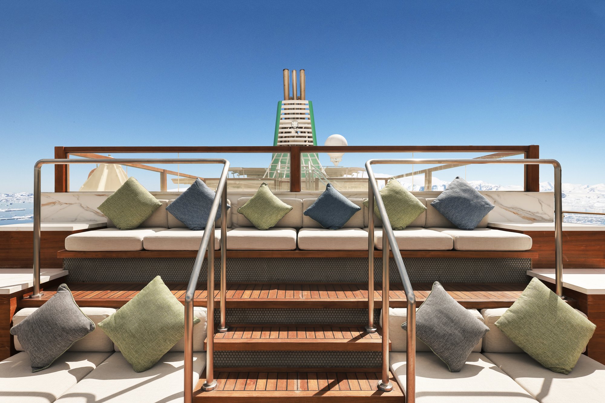 Luxury Deck Lounge with Arctic Views An elegant outdoor deck lounge area featuring modern seating with cream cushions and green accent pillows, arranged on wooden decking with metal frame structures. A distinctive funnel structure with three gold stacks rises prominently in the background, with snow-covered mountains and icy waters visible beyond the protective railings, suggesting an Arctic or polar cruise destination.
