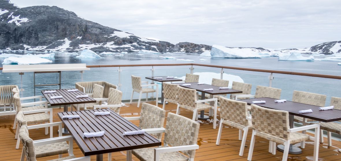 Luxury Dining Deck with Arctic Glacier Views An elegant outdoor dining and lounge area aboard a cruise ship overlooking a pristine Arctic landscape with icebergs, glaciers, and snow-covered mountains. Multiple dining tables with woven cream-colored chairs are arranged on a wooden deck behind a glass railing, offering panoramic views of the icy waters and mountainous terrain.