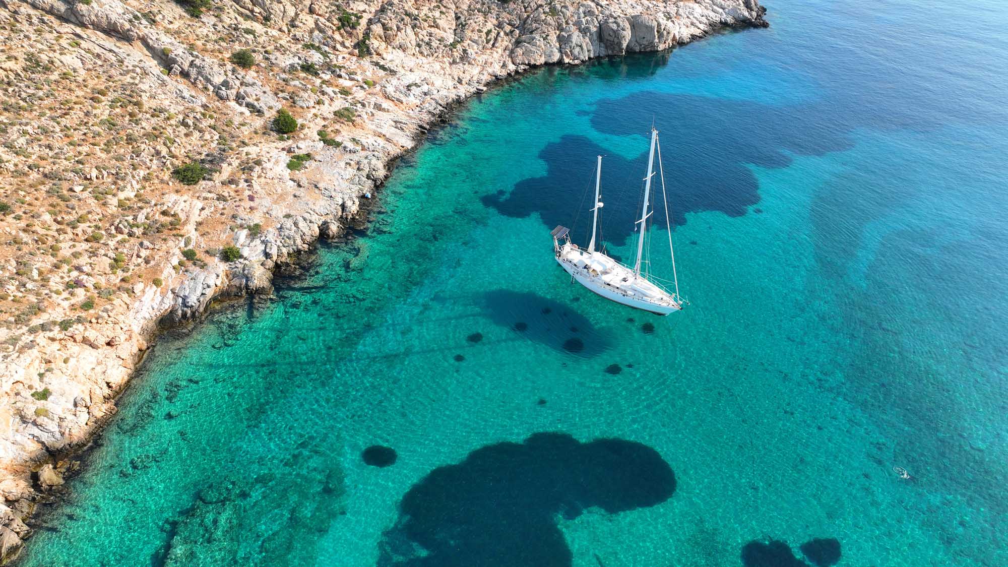 An aerial drone photograph of a white sailing yacht anchored in crystal-clear turquoise waters beside dramatic white limestone cliffs in Ornos Bay, Mykonos. The pristine Mediterranean seascape showcases the boat's reflection in the transparent water, with dark rocky outcrops visible beneath the surface.