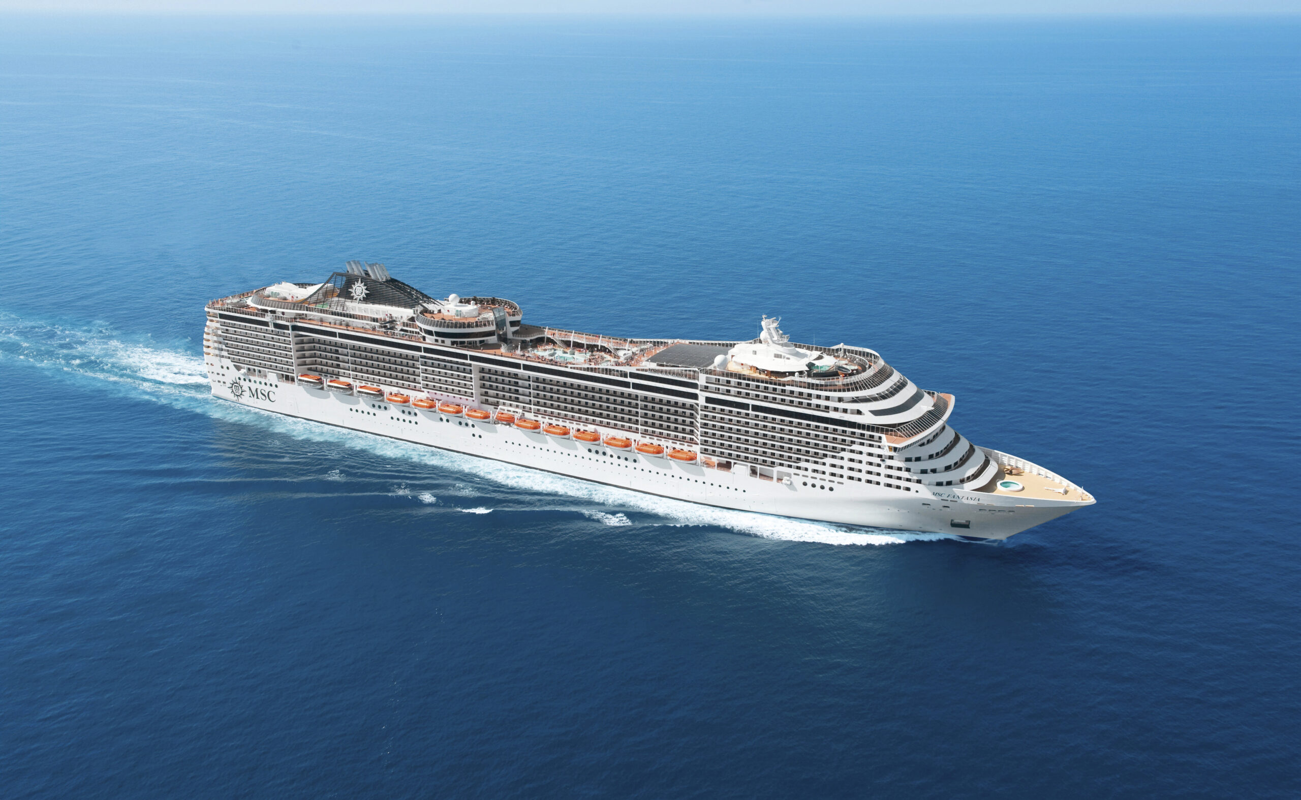 MSC Cruise Ship in Open Ocean A modern large cruise ship with distinctive MSC branding visible on the hull, sailing through calm blue waters. The vessel features multiple decks with orange and white exterior trim, characteristic of contemporary MSC fleet design, photographed from an aerial perspective on a clear day.