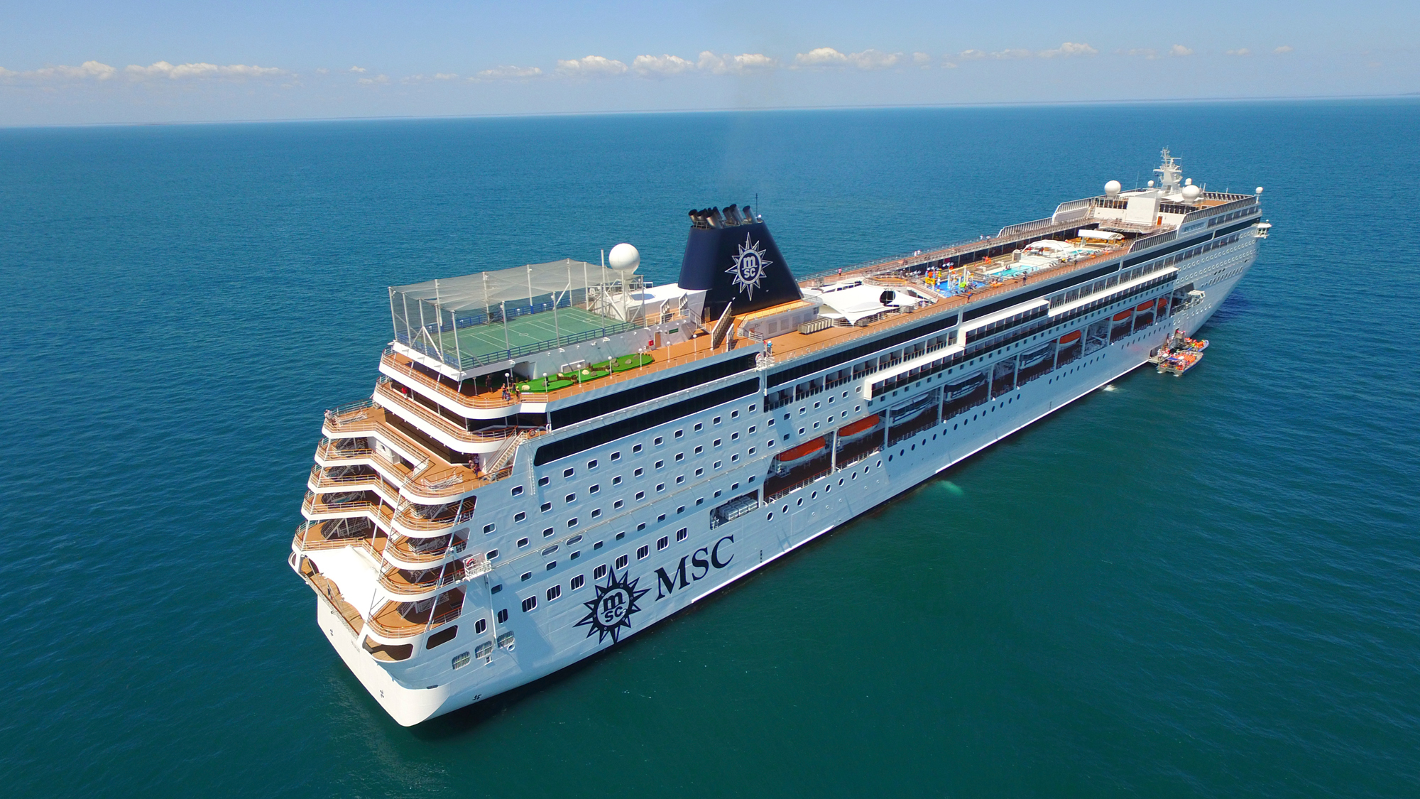 MSC Sinfonia Cruise Ship at Sea A large modern cruise ship with distinctive MSC branding and livery anchored in clear turquoise waters. The vessel features multiple passenger decks with balconies, a sports court on the upper deck, and the characteristic MSC funnel with wheel logo, photographed from an aerial perspective under clear skies.