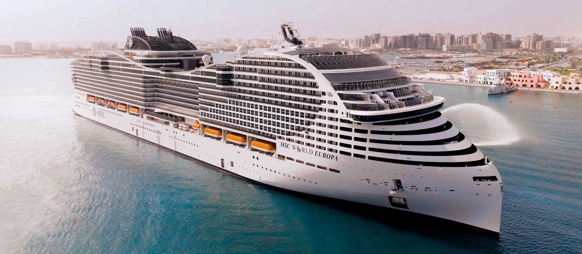 A magnificent modern cruise ship, the MSC World Europa, docked in a turquoise Mediterranean port with a coastal cityscape visible in the background. The vessel showcases distinctive contemporary design with tiered black and white balconies, multiple decks, and modern amenities characteristic of next-generation cruise ships.
