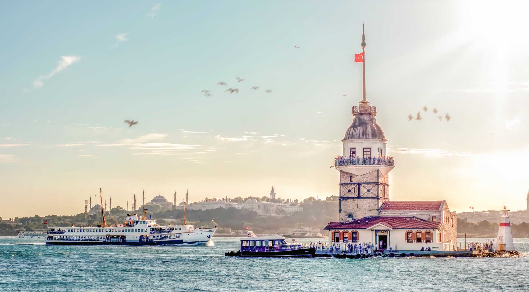 A scenic view of Istanbul's iconic Maiden's Tower (Kız Kulesi) in the Bosphorus Strait, with a blue and white passenger ferry approaching the tower and the cityscape of Istanbul visible in the background under a clear sky. The historic lighthouse-style tower features a domed top with Turkish flag, surrounded by water taxis and sightseeing vessels.