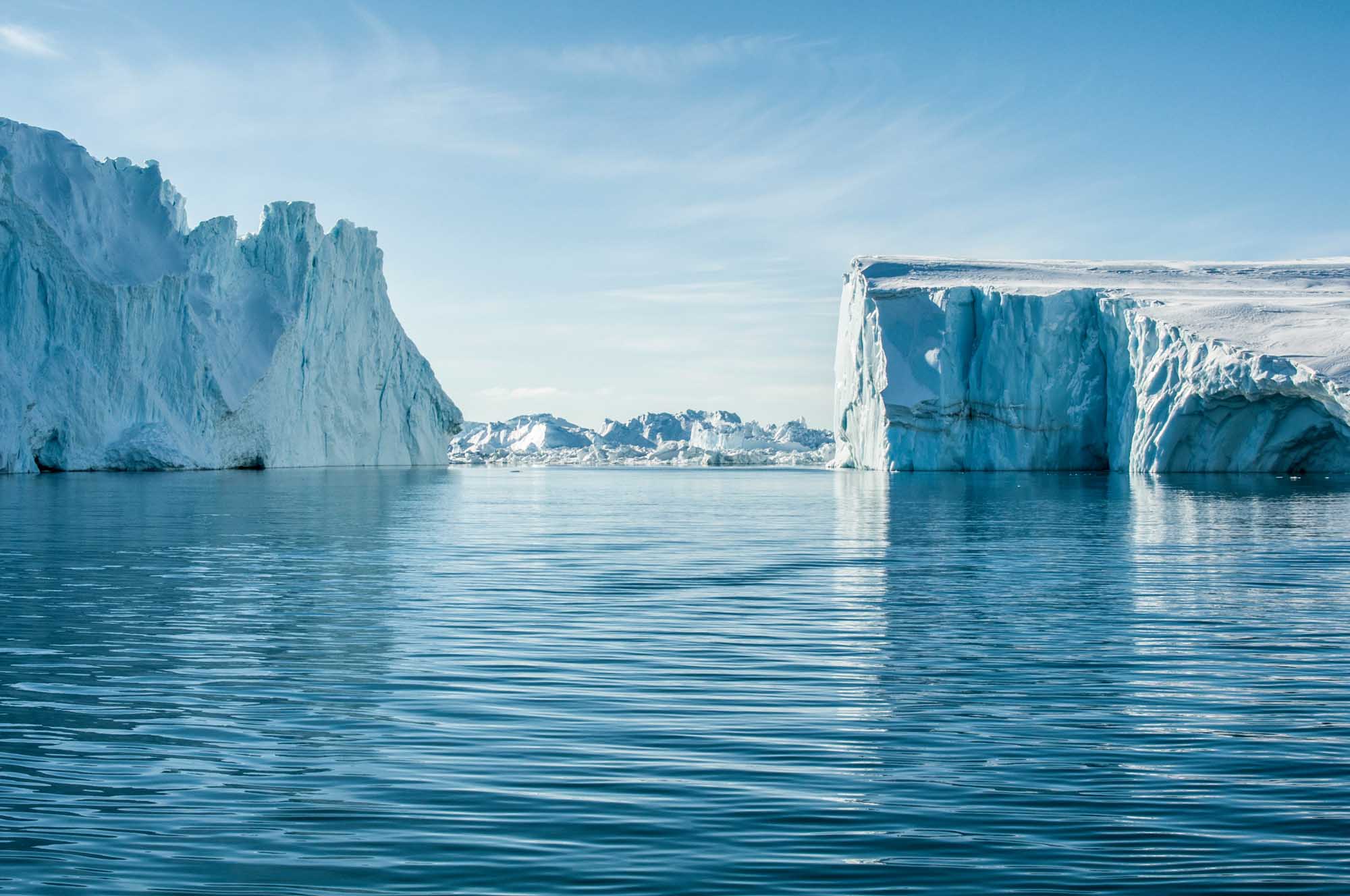 Majestic Icebergs in Greenlandic Waters Towering white and blue icebergs float in calm turquoise Arctic waters, with a small Greenlandic settlement visible in the distance across the bay. The dramatic ice formations showcase the raw beauty of Greenland's glacial landscape under clear skies.
