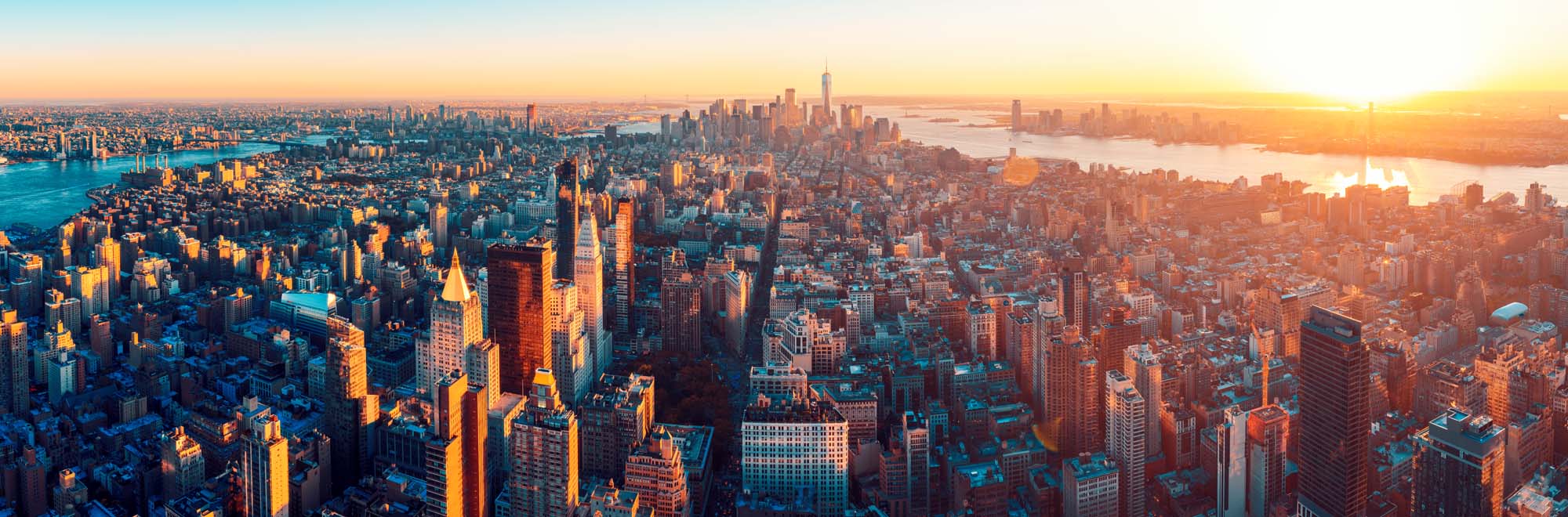 An expansive aerial panoramic view of Manhattan captured during golden hour, showcasing the iconic skyline with illuminated skyscrapers, the East River, Hudson River, and surrounding boroughs bathed in warm sunset light. The image captures the scale and density of New York City's architecture against a luminous golden sky.