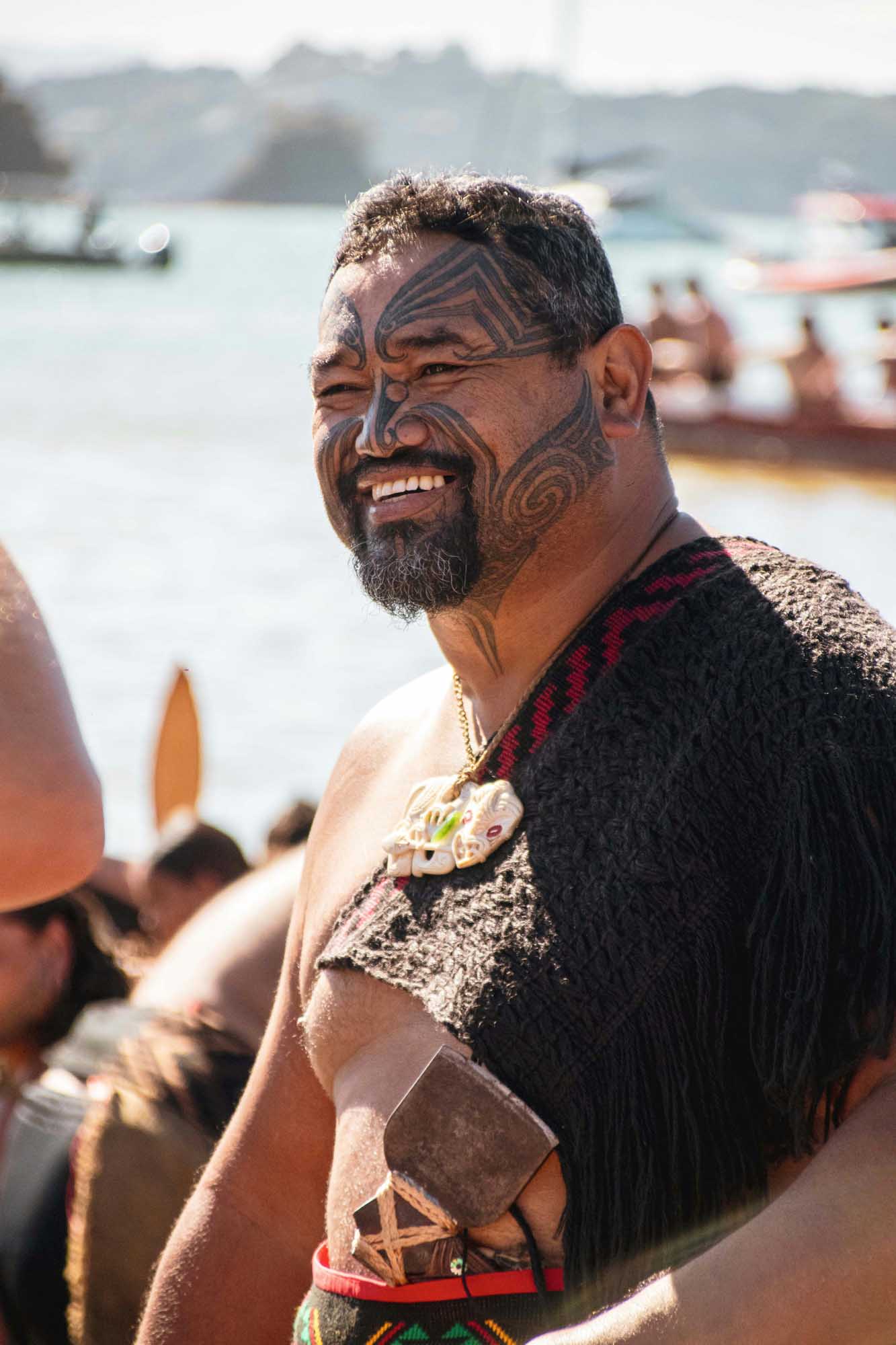 A man with traditional Maori ta moko facial tattoos smiles at the camera while standing near a waterfront with boats and hills in the background. He wears traditional Maori garments including a woven cloak and ornamental pendant, representing indigenous New Zealand culture.