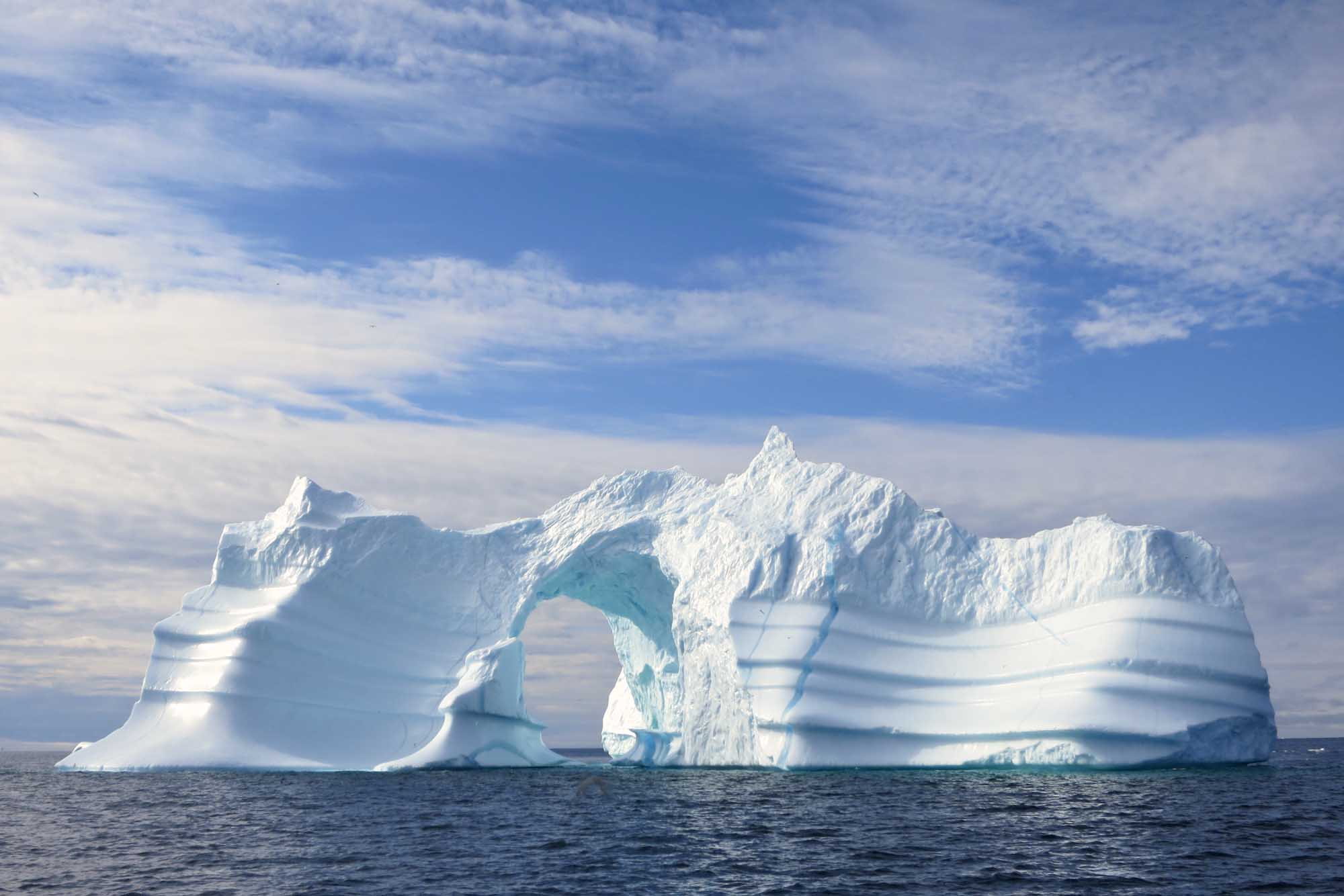 A striking natural arch iceberg floats in the dark waters of Greenland, featuring brilliant white ice sculpted by wind and waves into a distinctive tunnel-like formation. The dramatic ice formation is photographed against a partly cloudy blue sky, showcasing the raw beauty of Arctic glacial landscapes.