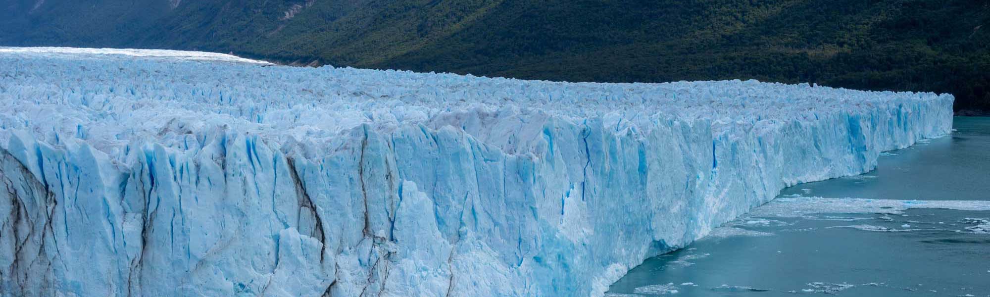 Massive Glacier Face with Turquoise Waters A dramatic view of a massive glacier terminus with striking blue-white ice formations rising from turquoise glacial meltwater, backed by dense forested mountains typical of Patagonia's stunning alpine landscape.