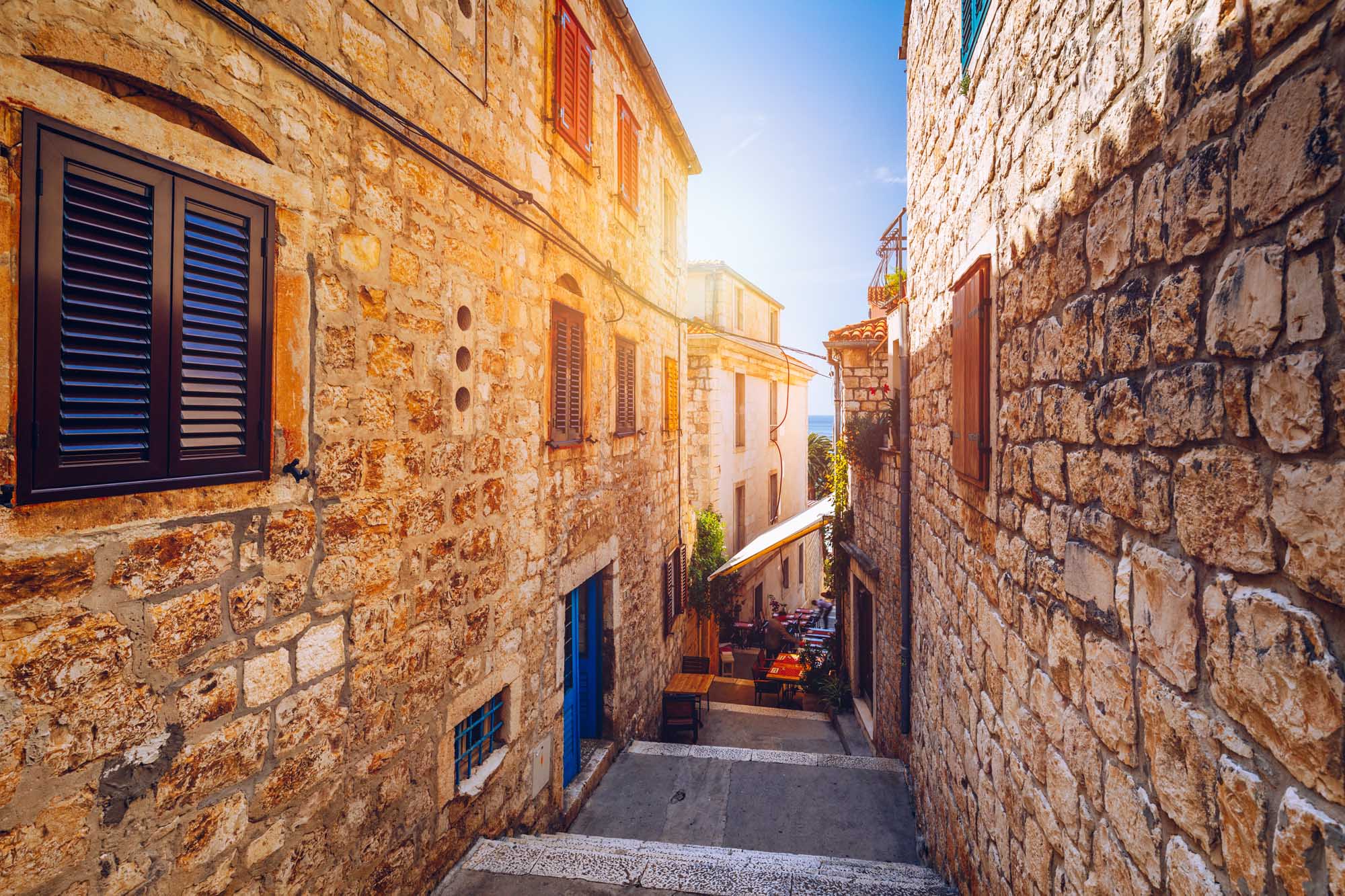 Medieval Stone Street in Hvar Town, Croatia A picturesque narrow alleyway in the historic town of Hvar on the island of Hvar, Dalmatia, Croatia. The scene features traditional Mediterranean stone architecture with terracotta and cream-colored buildings, wooden shutters in warm tones, and a charming pedestrian walkway lined with local establishments and sea views in the distance under clear blue skies.