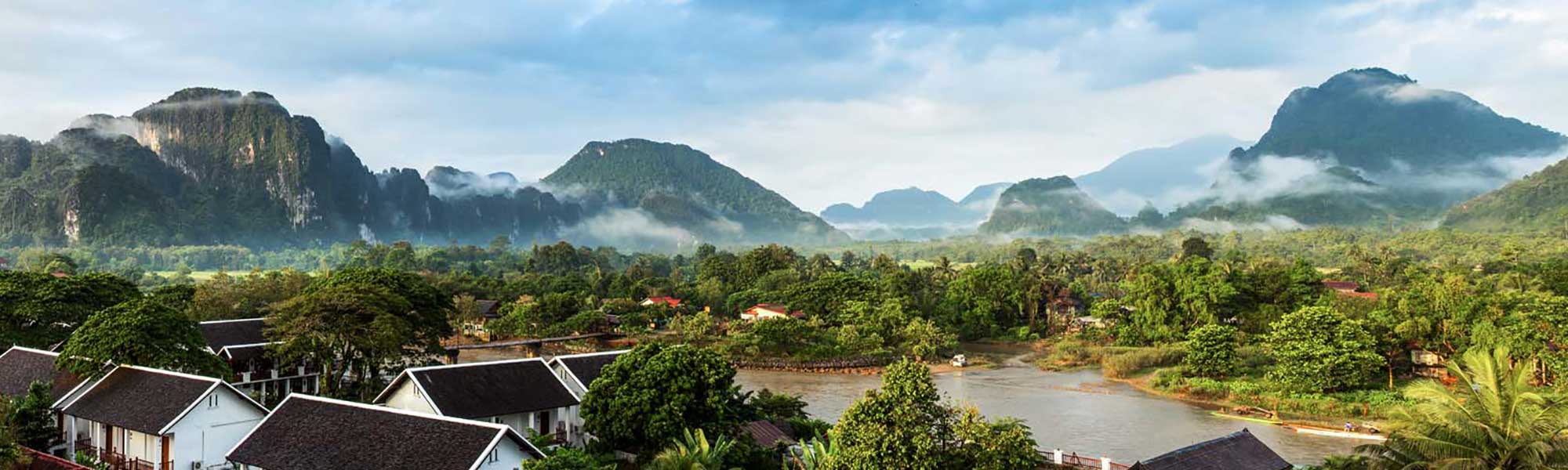 Mekong River Valley Landscape with Limestone Karsts A scenic panoramic view of the Mekong River valley in Laos, featuring dramatic limestone karst mountains shrouded in morning mist, traditional riverside buildings with dark tile roofs, lush tropical vegetation, and the calm waters of the Mekong River in the foreground.