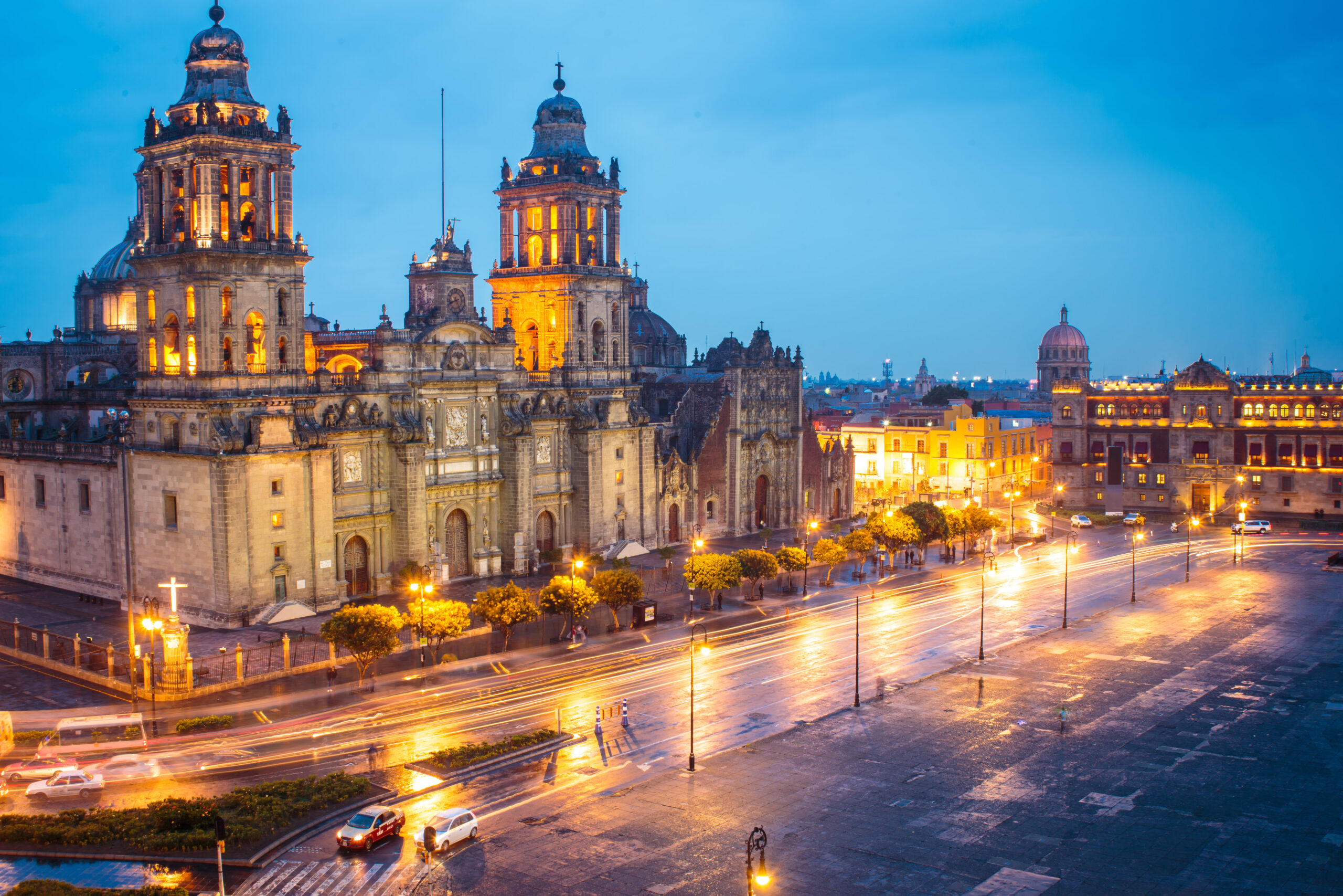 The stunning Mexico City Metropolitan Cathedral illuminated against a twilight blue sky, featuring twin baroque towers with golden lighting. The historic plaza below shows light trails from evening traffic, with colonial architecture visible throughout the cityscape.