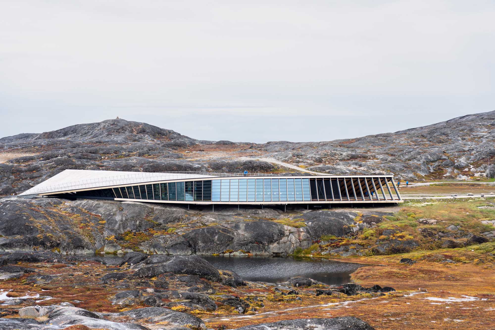 Modern Arctic Research Station in Greenland A striking contemporary building with a curved white roof and extensive blue-tinted glass panels sits atop rocky Arctic terrain in Greenland. The structure's sleek design contrasts dramatically with the barren, moss-covered landscape and distant mountains, exemplifying innovative architecture in one of the world's most remote regions.