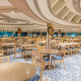 Modern Cruise Ship Lido Dining Area A spacious and elegantly designed lido dining area aboard a cruise ship, featuring multiple round tables with wooden chairs, blue patterned flooring, and an ornate curved ceiling with modern architectural details and recessed lighting. The venue showcases warm wood tones and extensive windows along the perimeter, creating a bright and inviting atmosphere for dining and socializing.