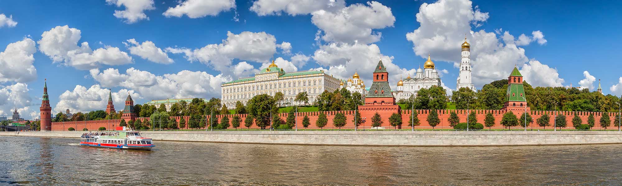Moscow Kremlin and River Cruise A scenic view of the iconic Moscow Kremlin fortress complex along the Moskva River, featuring its distinctive red brick walls, golden-domed cathedrals, and government buildings. A colorful tour riverboat passes in the foreground under clear skies with white clouds.