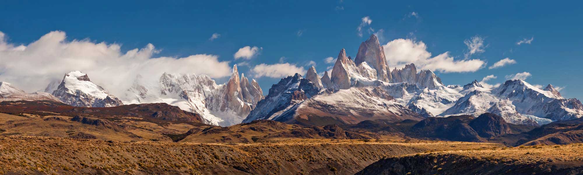 Mount Fitz Roy and Patagonian Peaks A dramatic panoramic vista of Mount Fitz Roy and the surrounding Patagonian mountain range in Argentina, featuring distinctive granite spires snow-capped peaks rising above arid, golden-brown steppe landscape under a brilliant blue sky with scattered white clouds.