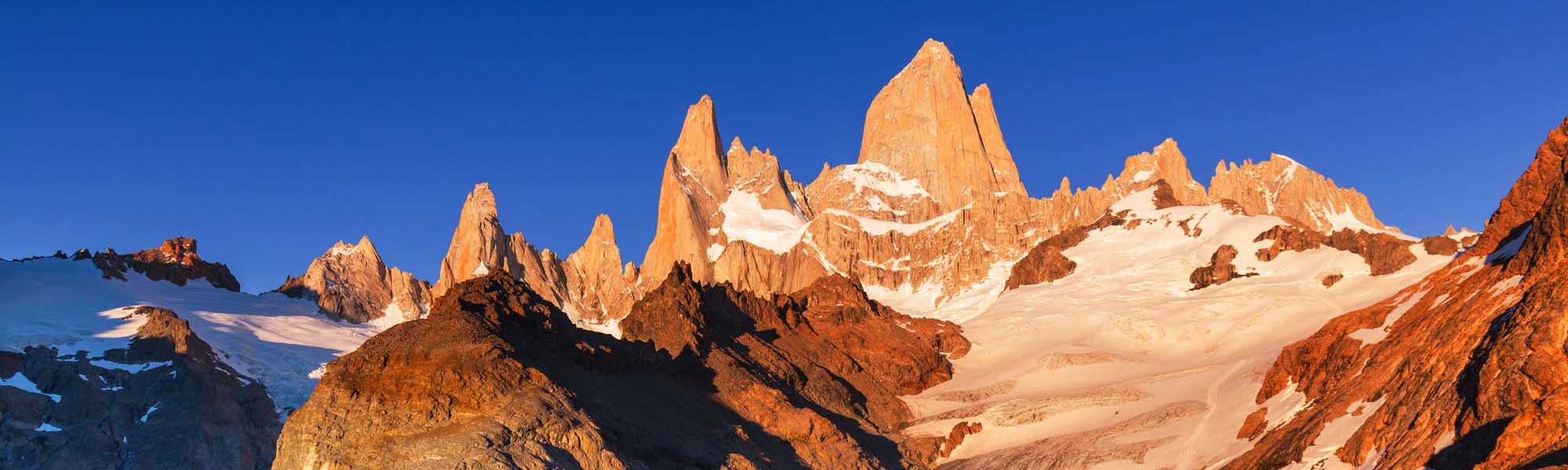 Mount Fitz Roy at Sunrise, Patagonia The iconic granite peaks of Mount Fitz Roy rise dramatically against a brilliant blue sky, their distinctive spires illuminated by golden morning light. Snow-covered glaciers and scree slopes surround the mountain in this stunning view of one of South America's most spectacular natural landmarks.