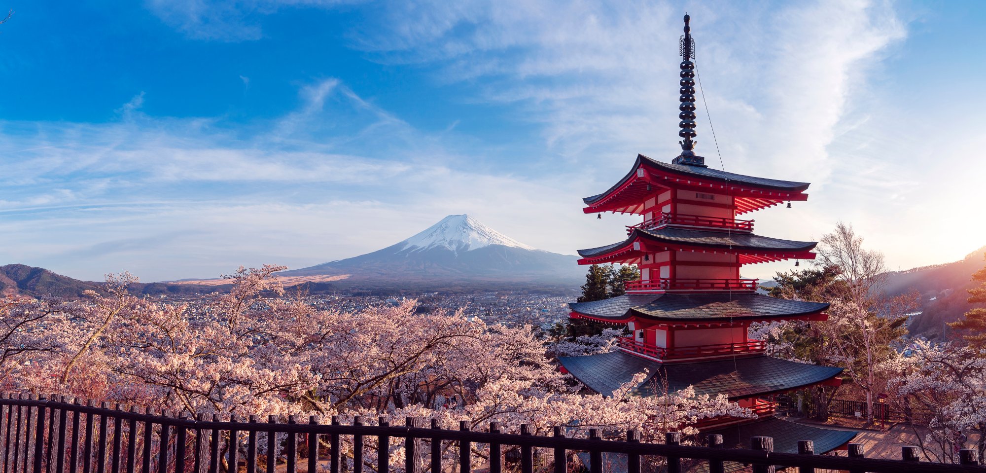 A stunning scenic view of a traditional red Japanese pagoda framed by cherry blossom trees in full bloom, with snow-capped Mount Fuji visible in the distance across the landscape. The composition captures the iconic beauty of Japan during spring with the pagoda's distinctive architecture prominent in the foreground.
