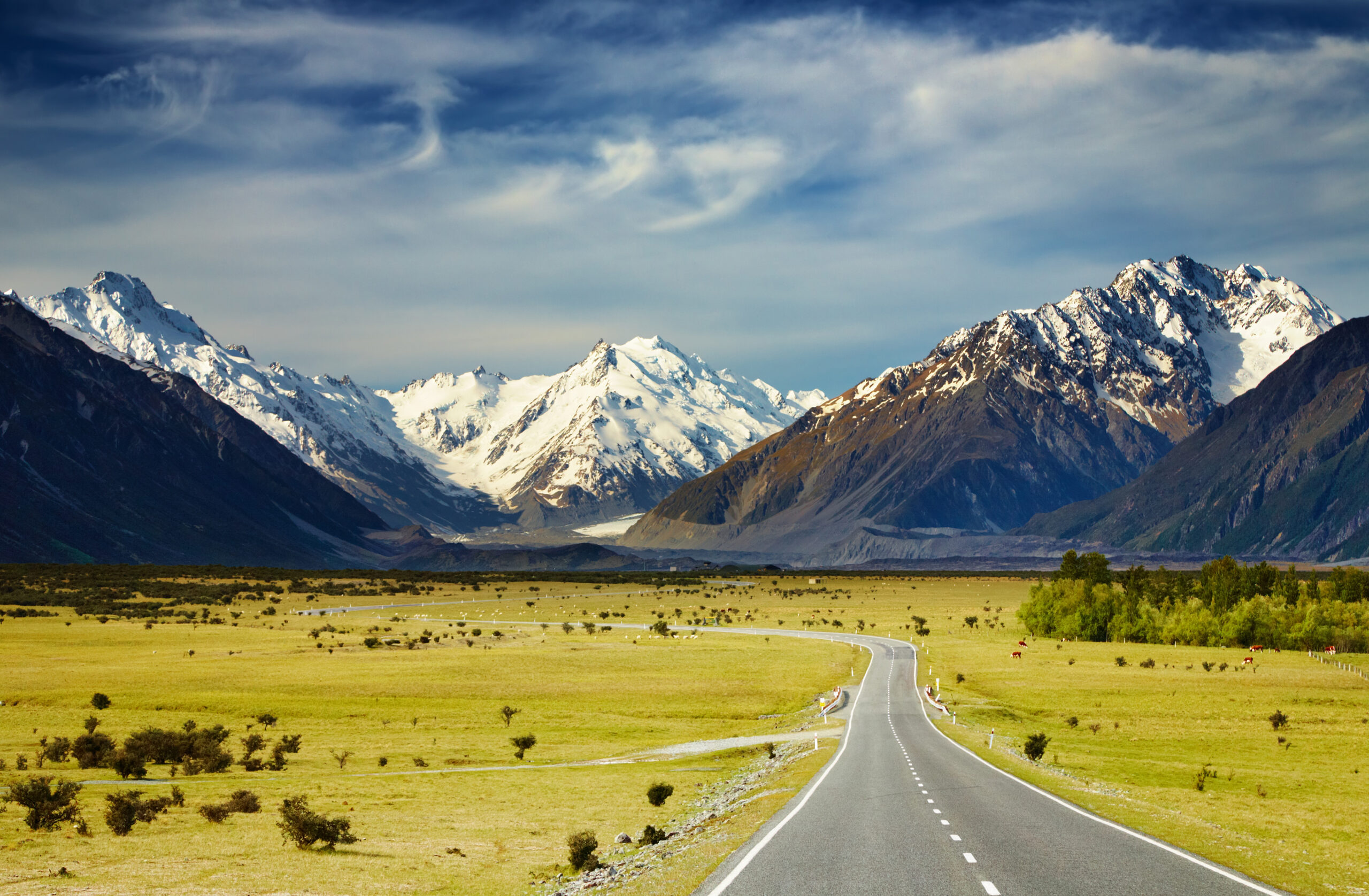 Mountain Road Through Alpine Valley A straight asphalt highway stretches through a verdant alpine valley dotted with sparse vegetation, leading toward a dramatic backdrop of snow-capped mountain peaks under a partly cloudy sky. The scene captures the raw beauty of high-altitude mountainous terrain with pristine landscape and clear road markings.