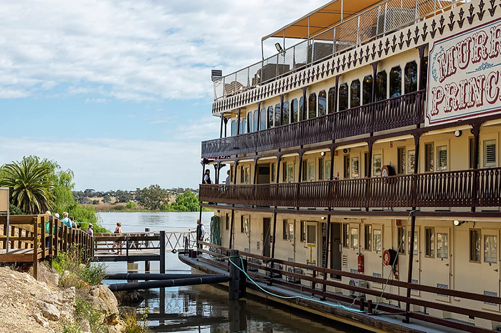 A three-deck paddlewheel riverboat with cream and brown colonial-style architecture docked along a peaceful river, featuring ornate railings and multiple levels of verandahs. The vessel displays classic Australian riverboat design with traditional paddle-wheel aesthetics and serves as a floating accommodation for river cruises.