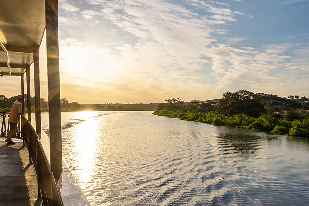 A scenic view of the Murray River at golden hour, captured from the deck of a riverboat. The sun reflects beautifully across the calm water, with lush green vegetation lining both riverbanks under a partly cloudy sky.