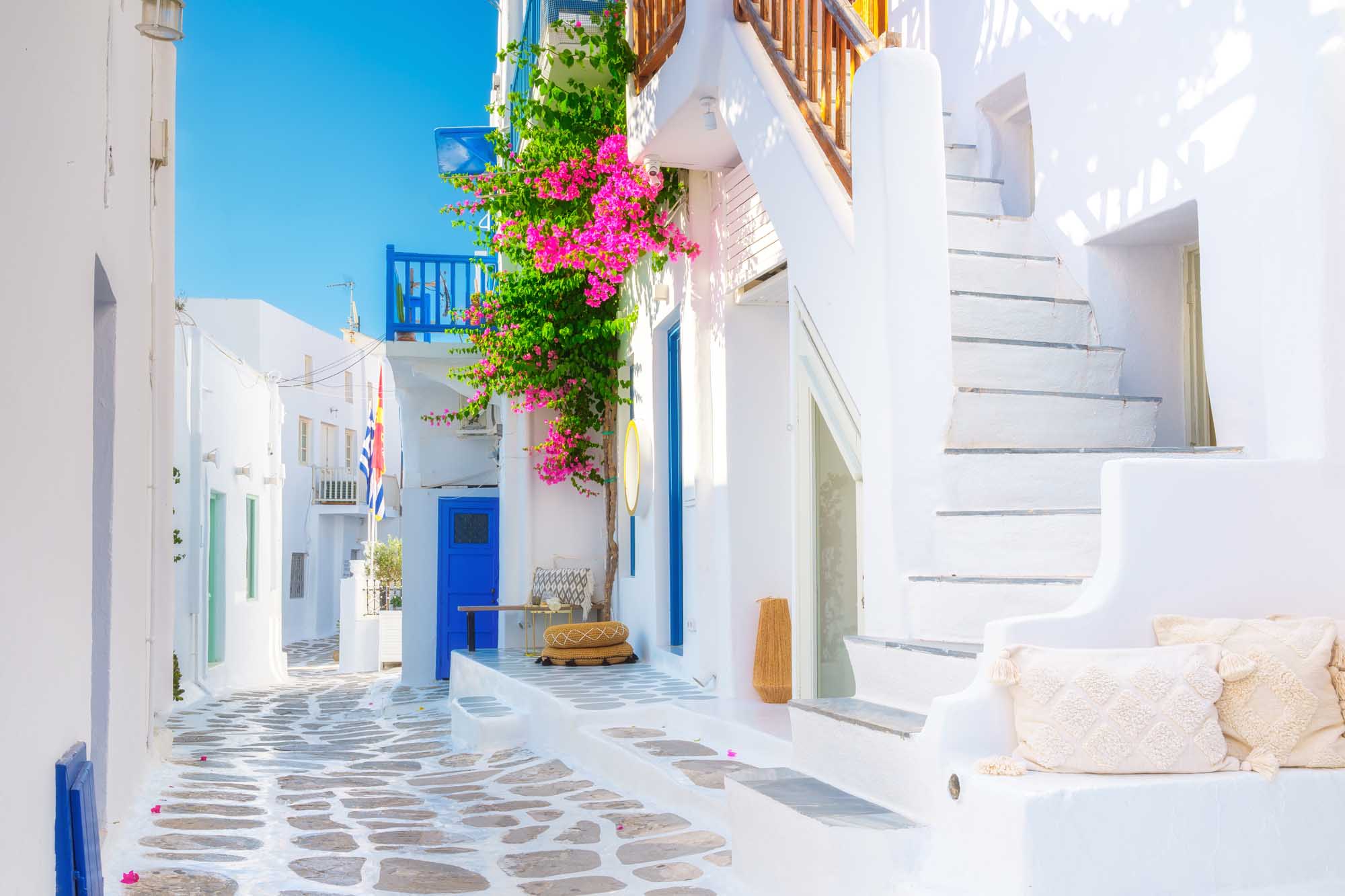 A charming narrow alleyway in Mykonos, Greece, featuring quintessential Cycladic architecture with whitewashed buildings, blue doors and shutters, and vibrant magenta bougainvillea cascading down a building. The traditional cobblestone street, bright Mediterranean sunlight, and picturesque Greek island ambiance make this an iconic travel destination scene.
