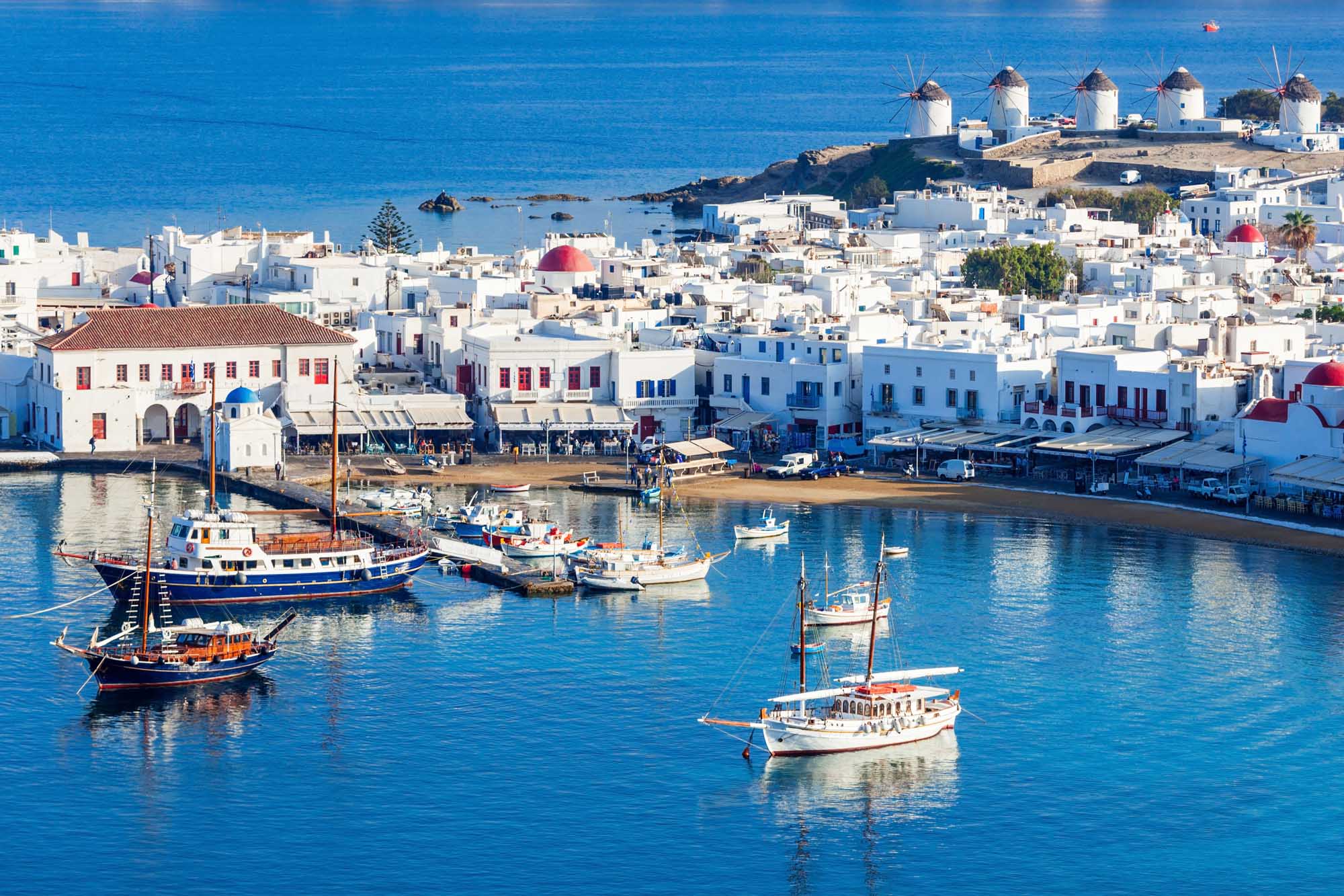 A picturesque view of Mykonos, Greece, featuring the iconic whitewashed windmills perched on a hillside overlooking the turquoise harbor filled with traditional fishing boats and sailboats. The charming Cycladic architecture with blue-domed roofs and white-washed buildings cascades down to the waterfront, creating a quintessential Mediterranean island scene.