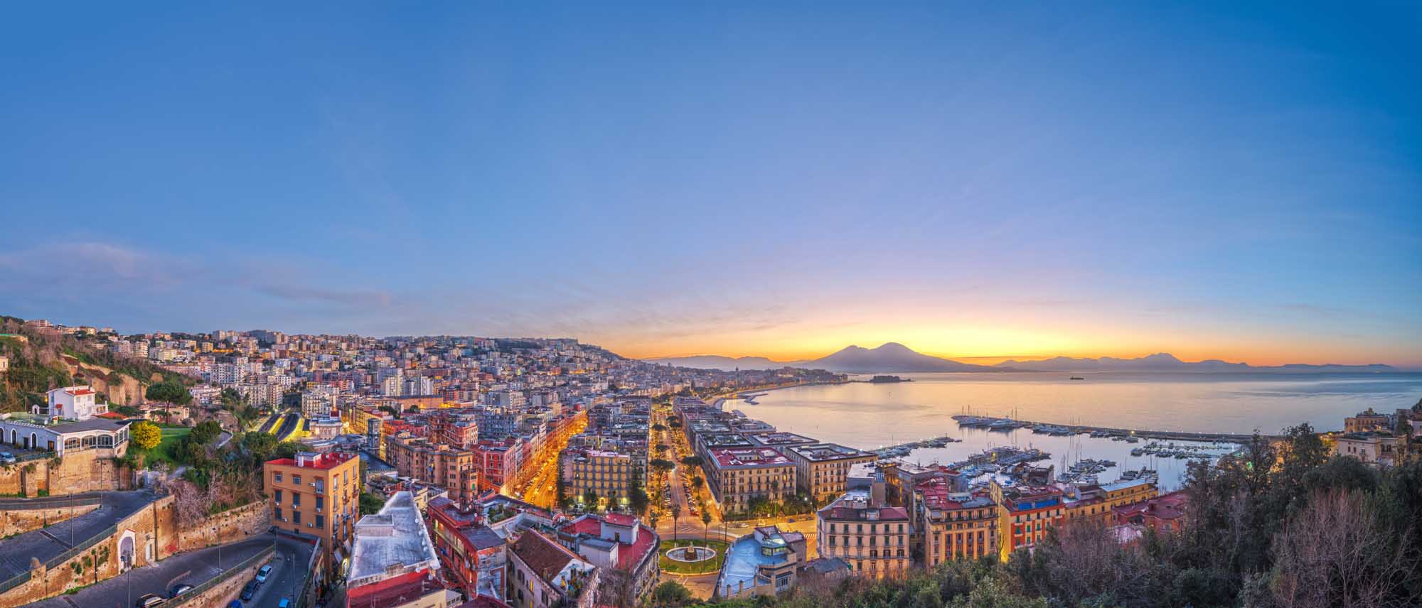 An expansive aerial view of Naples, Italy at sunrise, showcasing the densely-packed colorful buildings cascading down hillsides toward the Bay of Naples. Mount Vesuvius is visible on the horizon across the bay, silhouetted against the golden dawn sky, while numerous sailboats dot the harbor below.