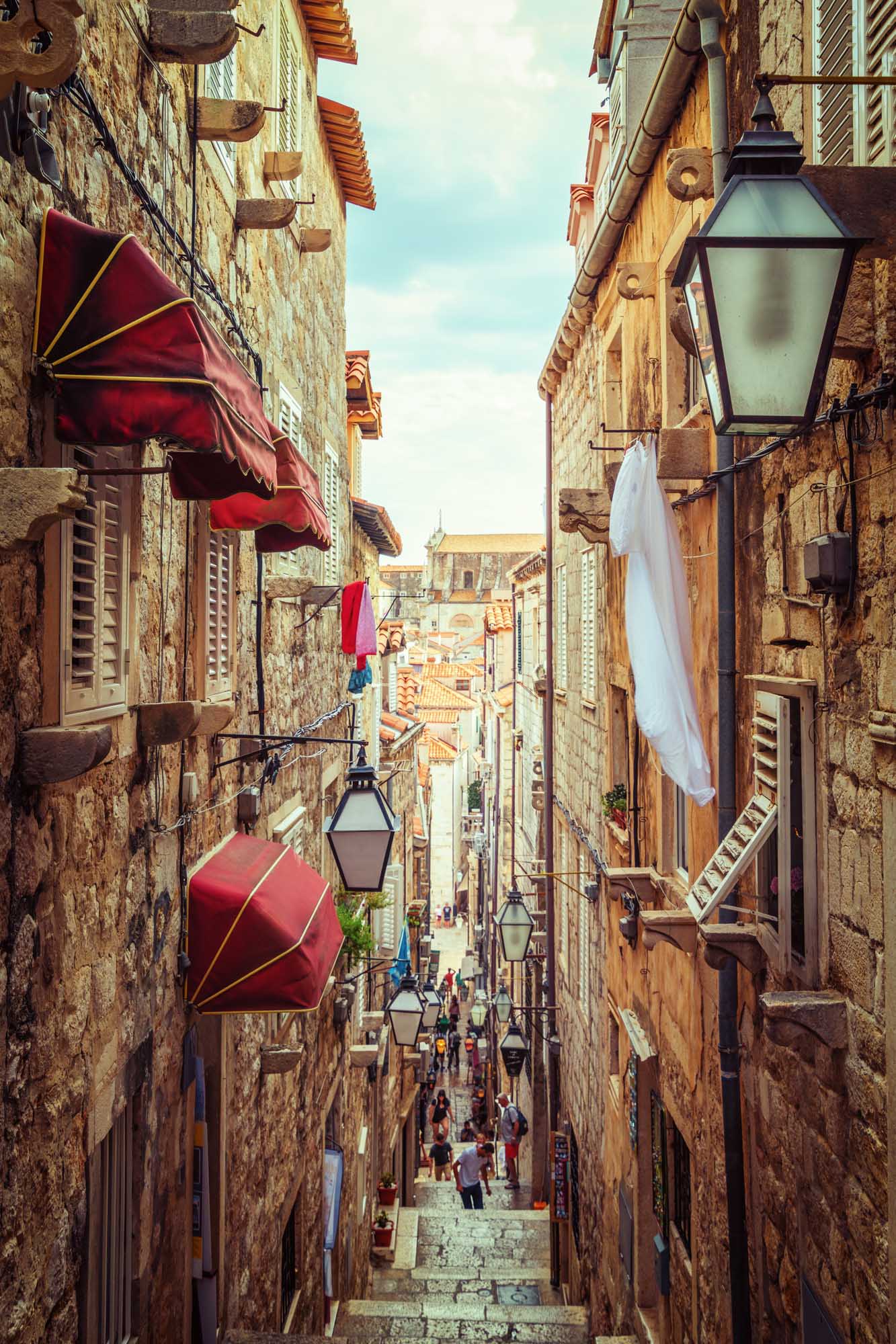 A picturesque narrow stone alley in Dubrovnik's UNESCO-listed Old Town, lined with traditional architecture, red awnings, vintage lanterns, and white laundry hanging between buildings. Tourists stroll through the charming medieval street with terracotta roofs visible in the background under a clear blue sky.