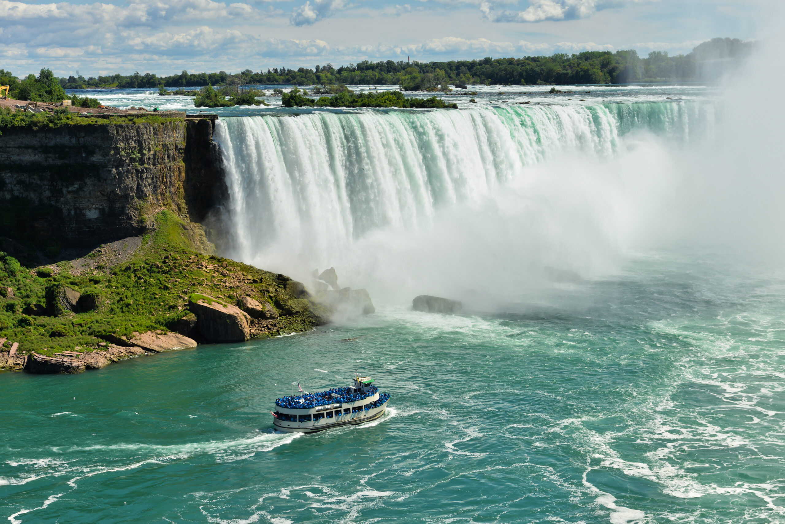 Niagara Falls with Tourist Boat Tour A scenic view of Niagara Falls' Horseshoe Falls with a passenger tour boat navigating the turquoise waters below, surrounded by mist and lush greenery on the Canadian side. The iconic natural wonder showcases the massive cascade of water with a blue-sky backdrop and forested landscape.