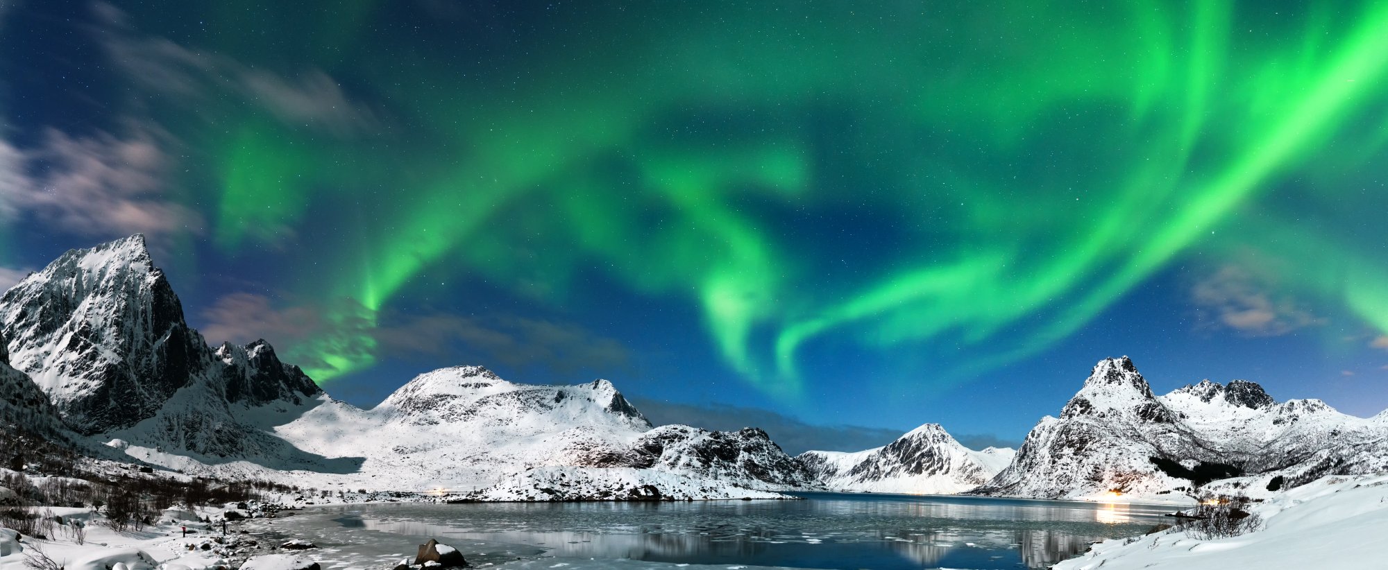 A breathtaking Arctic landscape featuring vibrant green aurora borealis dancing across the night sky above a serene frozen fjord, surrounded by majestic snow-covered peaks and dark rocky outcrops. The ethereal light display reflects on the calm water below, creating a stunning natural spectacle characteristic of polar regions.