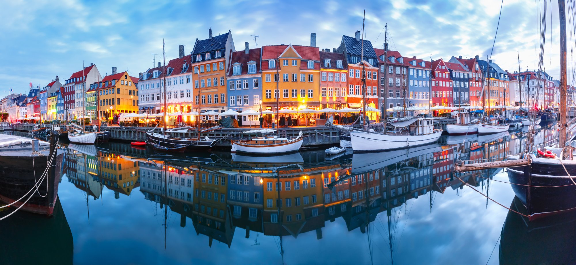 A stunning twilight view of Copenhagen's iconic Nyhavn waterfront, featuring a row of vibrant multi-colored 17th-century buildings reflected perfectly in the still harbor waters, with traditional sailing boats moored alongside the quay and warm evening lights illuminating the historic facades.