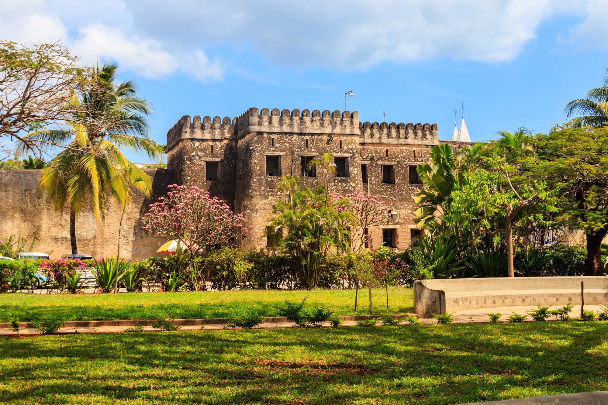 Old Fort in Stone Town, Zanzibar The historic Old Fort (Arab Fort), a centuries-old stone fortification in Stone Town, Zanzibar, is surrounded by lush tropical gardens with blooming pink flowers and palm trees. The massive crenellated walls and multiple windows showcase classic Arab-Swahili architecture, with a flag flying from the rooftop against a clear blue sky.