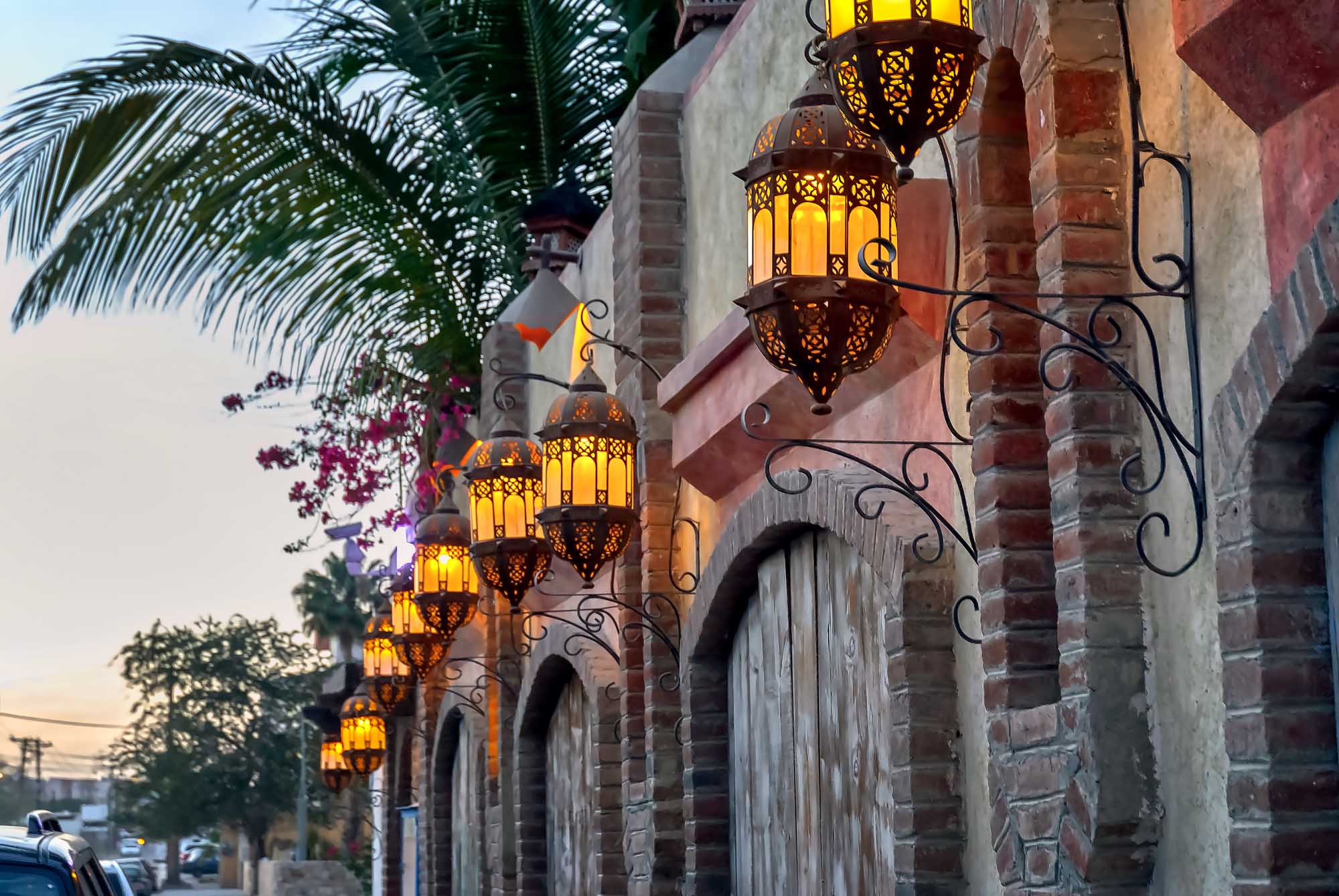 Decorative Moroccan-style hanging lanterns with intricate metalwork and golden glass illuminate the brick facade of a historic building in Cabo San Lucas at dusk. Vibrant bougainvillea flowers and a palm tree frame the architectural details, creating a warm, inviting ambiance typical of this popular coastal destination.