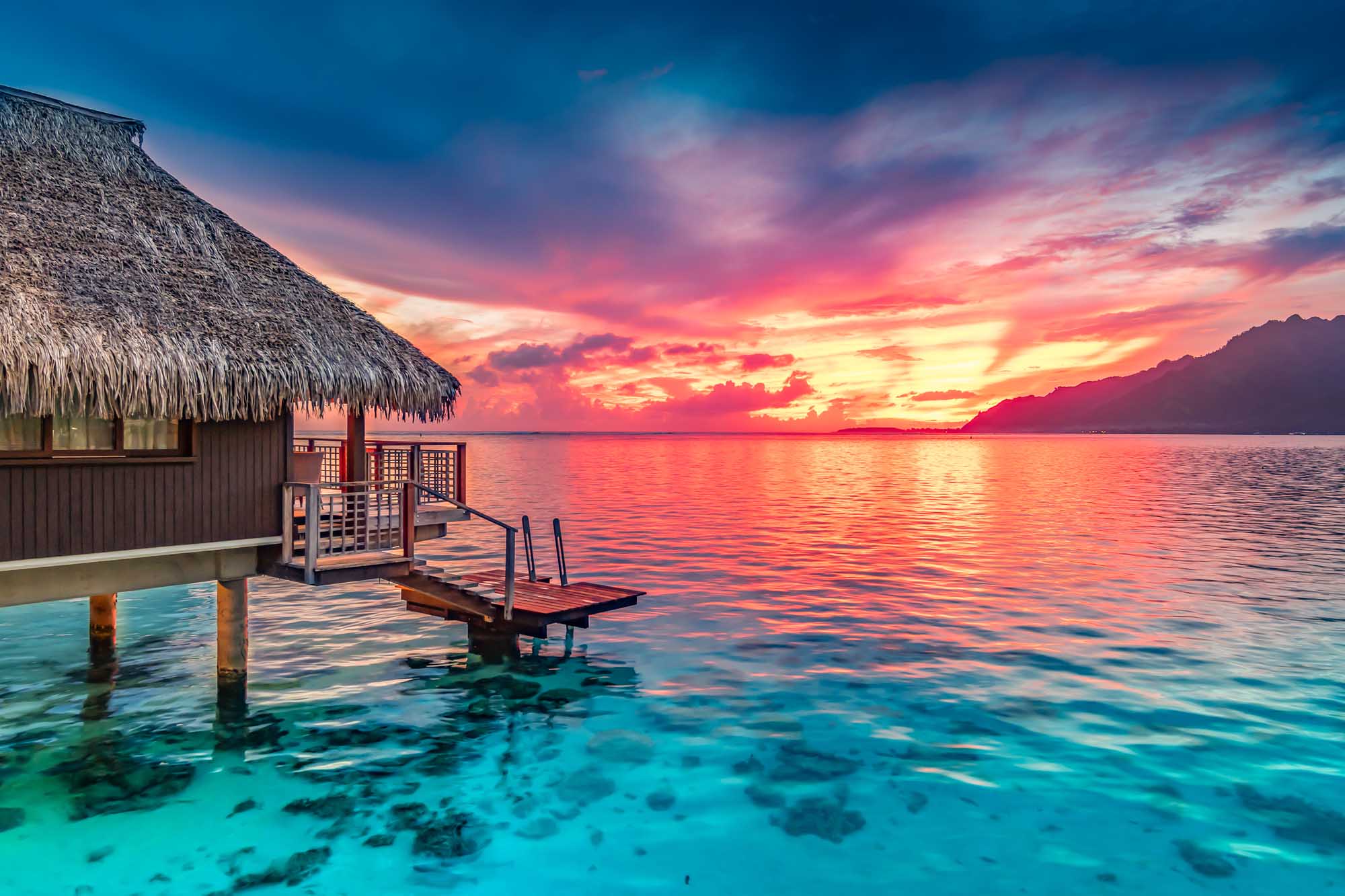 A stunning tropical sunset scene featuring a traditional thatched-roof overwater bungalow with wooden deck and jetty in crystal-clear turquoise waters. The dramatic sky displays vibrant hues of pink, orange, and gold reflecting across the calm ocean, with distant volcanic mountains silhouetted on the horizon.