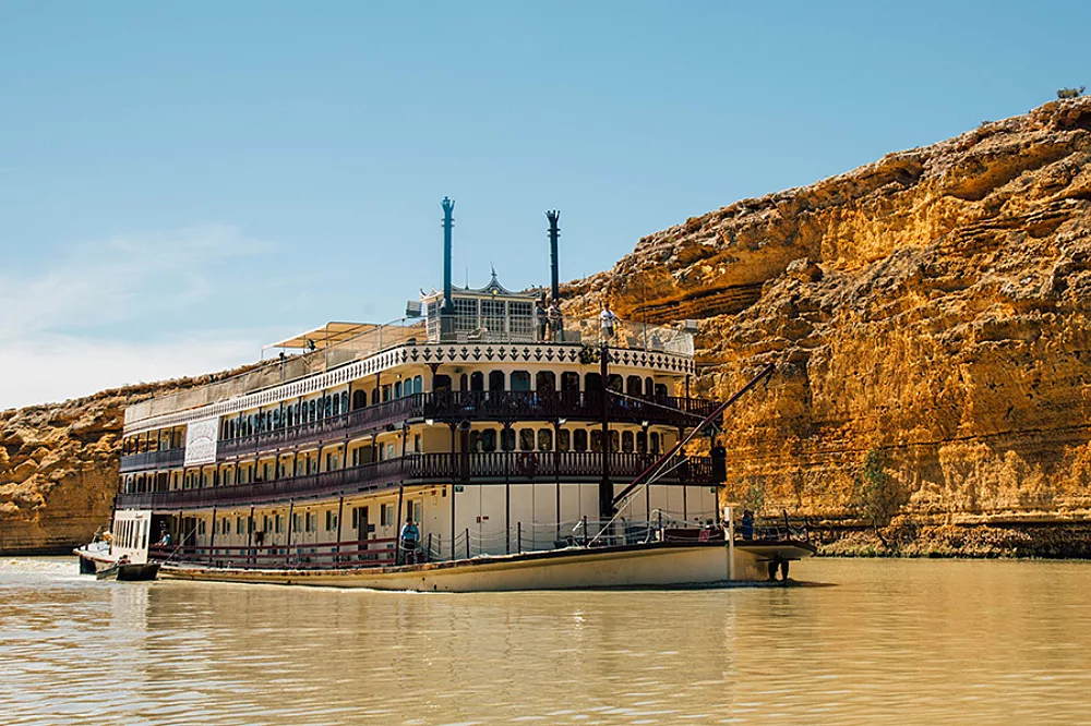 Paddlewheel Riverboat on Muddy River A classic multi-deck paddlewheel riverboat with distinctive twin smokestacks navigates a muddy river, with dramatic golden-hued limestone cliffs towering in the background. The vessel features traditional stern-wheel design with ornate white railings and multiple passenger decks typical of Mississippi River-style cruising.