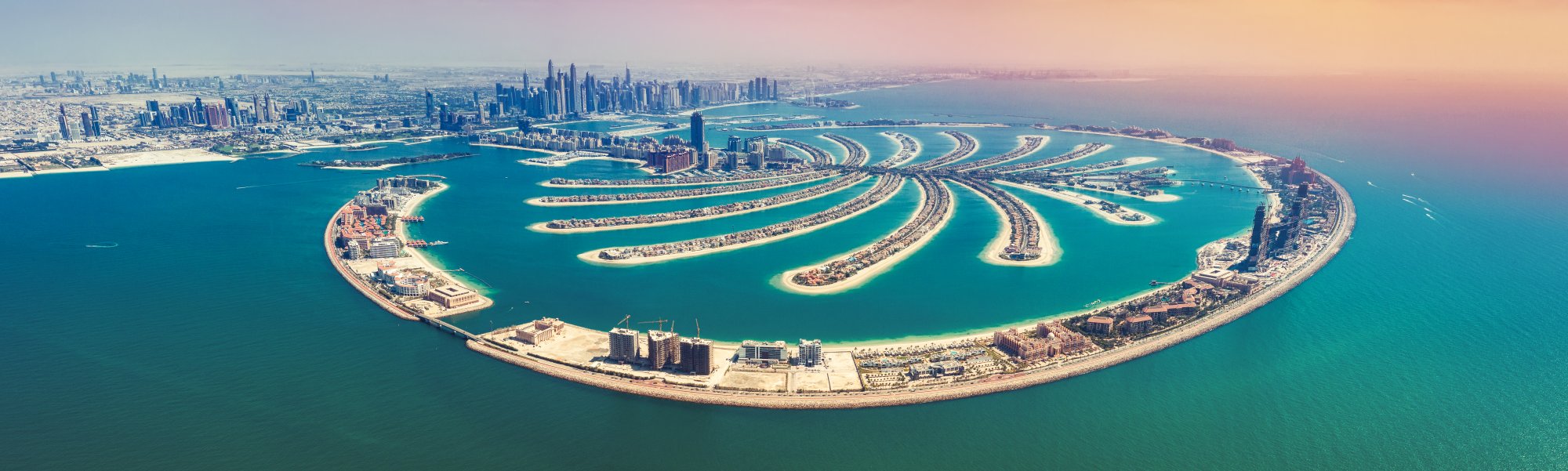 Palm Jumeirah Aerial View Dubai An aerial panoramic view of Palm Jumeirah, the iconic artificial palm-shaped island development in Dubai, United Arab Emirates. The image showcases the distinctive crescent-shaped waterfront with luxurious residential villas arranged in curved patterns, surrounded by turquoise Arabian Gulf waters, with the Dubai skyline and downtown cityscape visible in the background.