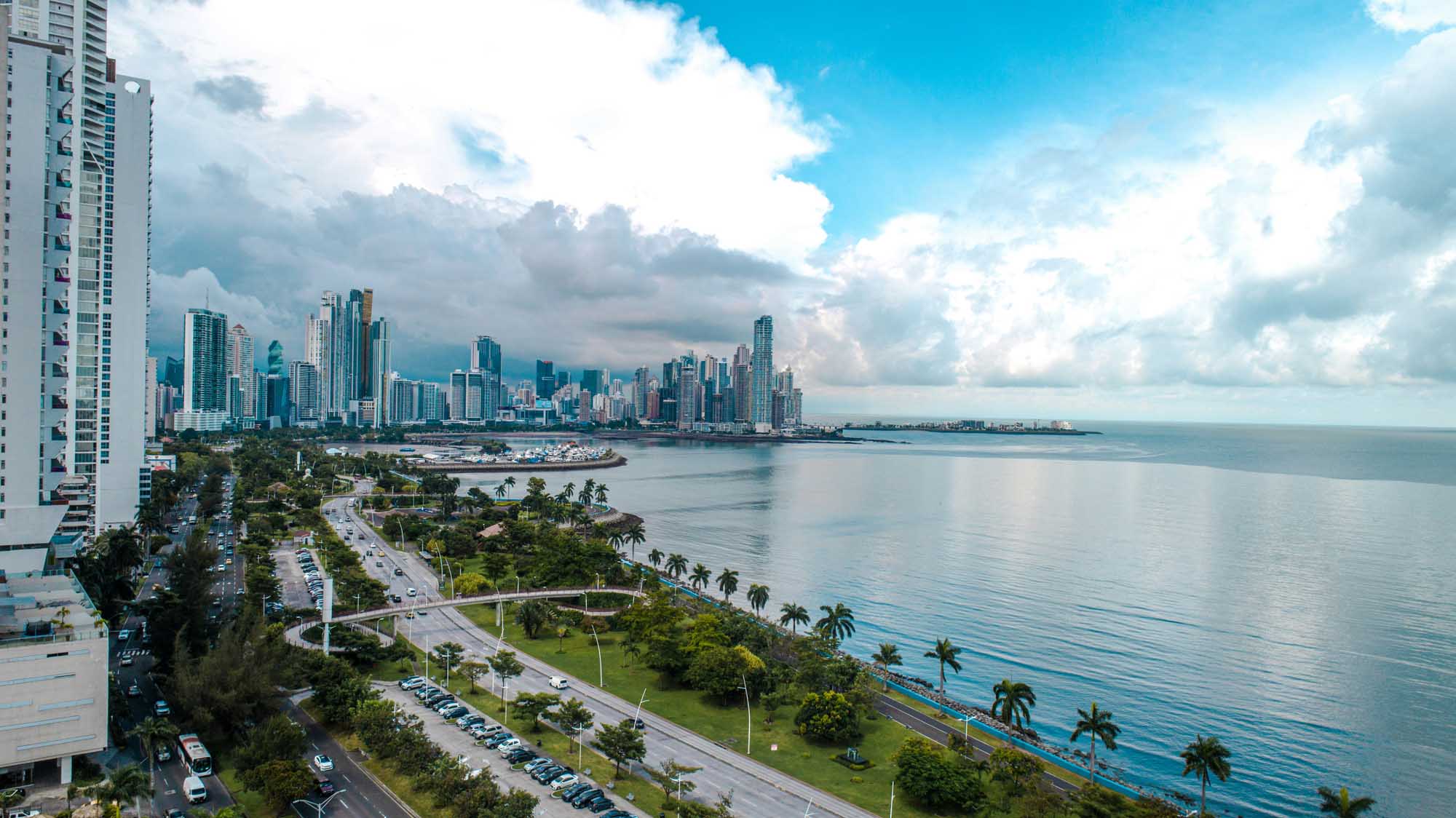 Panama City Skyline and Coastal Waters A scenic aerial view of Panama City's modern high-rise skyline along the Pacific coast, featuring contemporary residential and commercial towers, a manicured waterfront park with palm trees, and calm turquoise waters under a partly cloudy sky.