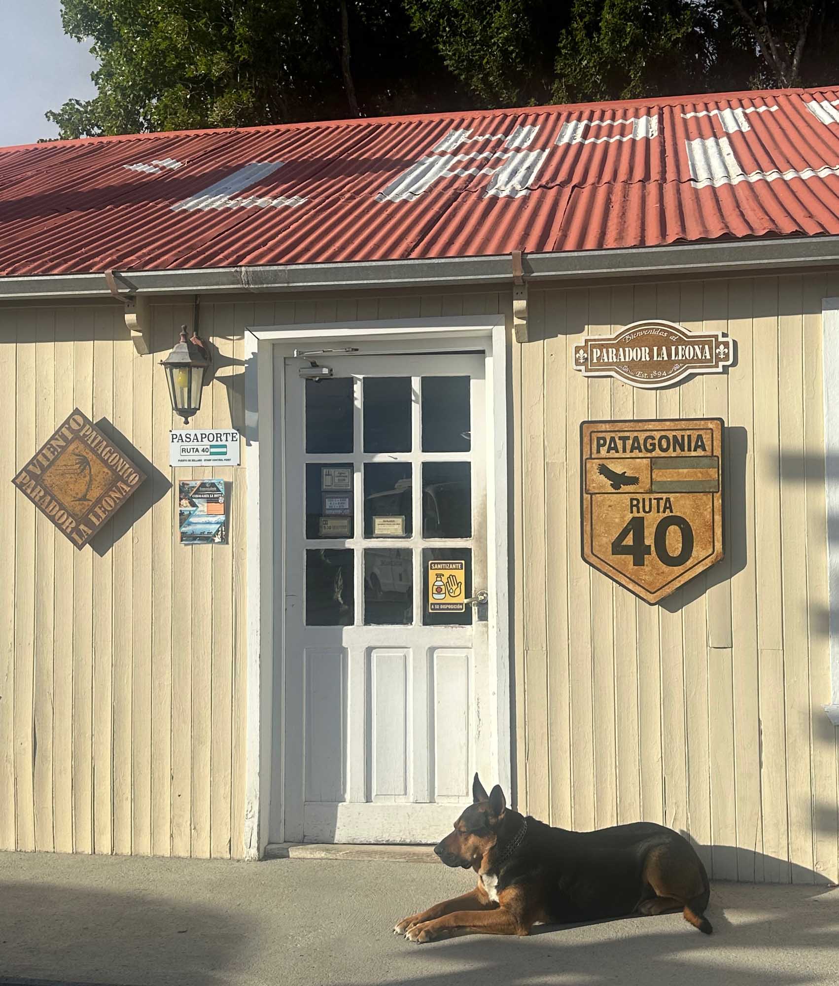 Parador La Leona Estancia, Patagonia Route 40 A charming rustic lodge building with cream-colored corrugated metal siding and a red metal roof, featuring vintage signage for 'Parador La Leona' and 'Patagonia Ruta 40'. A German Shepherd dog rests on the ground in front of the entrance, which displays traditional Patagonian hospitality aesthetic.