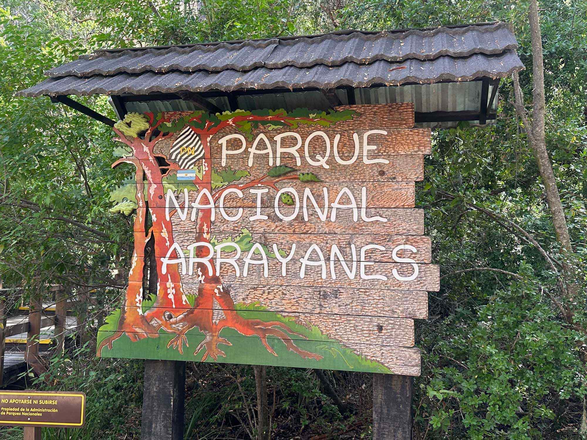 Parque Nacional Arroyanes National Park Entrance Sign A rustic wooden entrance sign for Parque Nacional Arroyanes (Arrayanes National Park) surrounded by lush green forest vegetation. The brick and wood structure features colorful indigenous-inspired artwork including a red figure and natural elements, with a weathered corrugated metal roof overhead.
