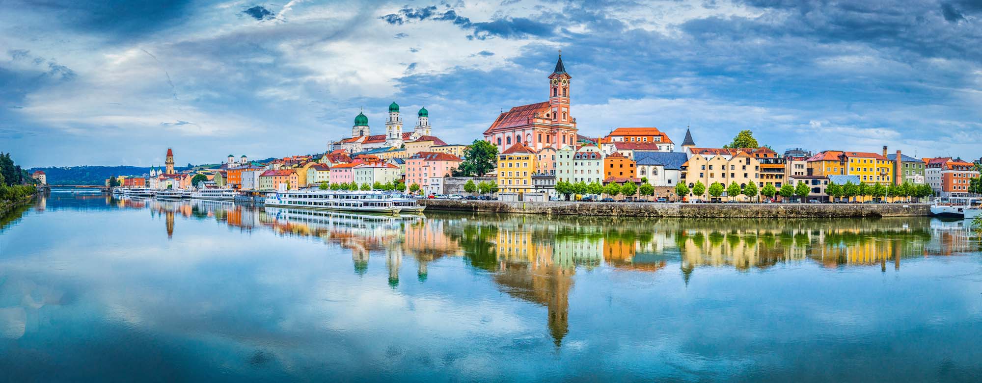 A panoramic view of the historic Bavarian city of Passau with its iconic colorful buildings and church spires reflected perfectly in the calm Danube River during golden hour. The picturesque riverside town showcases traditional Central European architecture with vibrant pastel-colored facades and a prominent red-roofed cathedral, creating a stunning mirror image on the water.