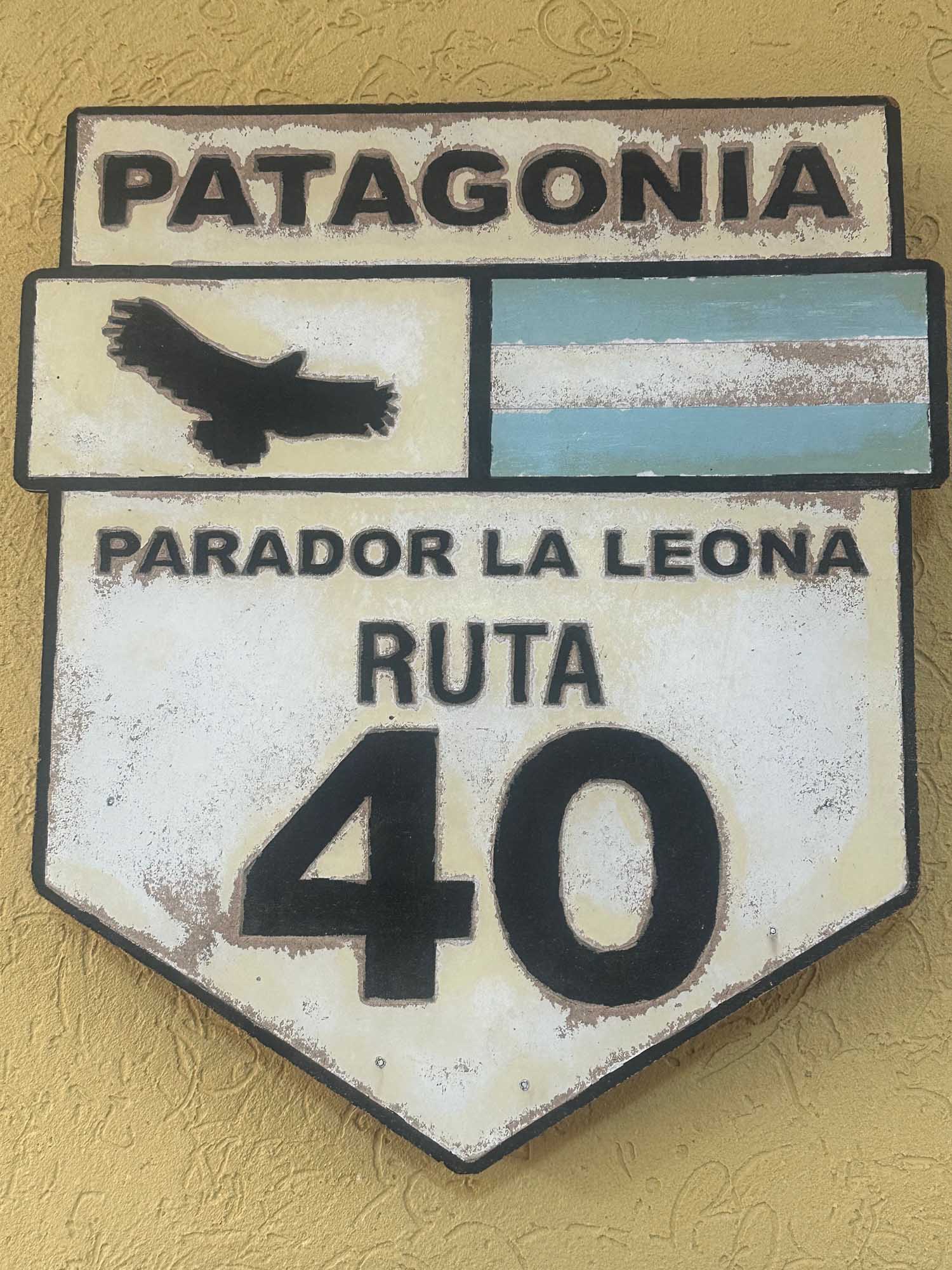 Patagonia Route 40 Directional Sign A weathered vintage-style road sign for Patagonia's Route 40 (Ruta 40), featuring the text 'PARADOR LA LEONA RUTA 40' with a silhouette of a condor and the Argentine flag stripes. The sign shows aged distressing and is mounted on a textured yellow wall, representing iconic South American highway signage.